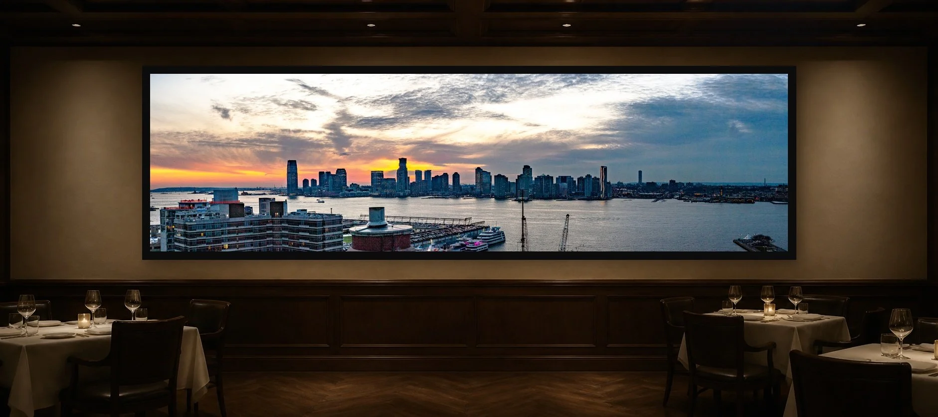 Skyline at Dusk displayed in a black frame within an intimate fine-dining room with plaster walls, dark wood, linen tables, no guests, and a clear central wall