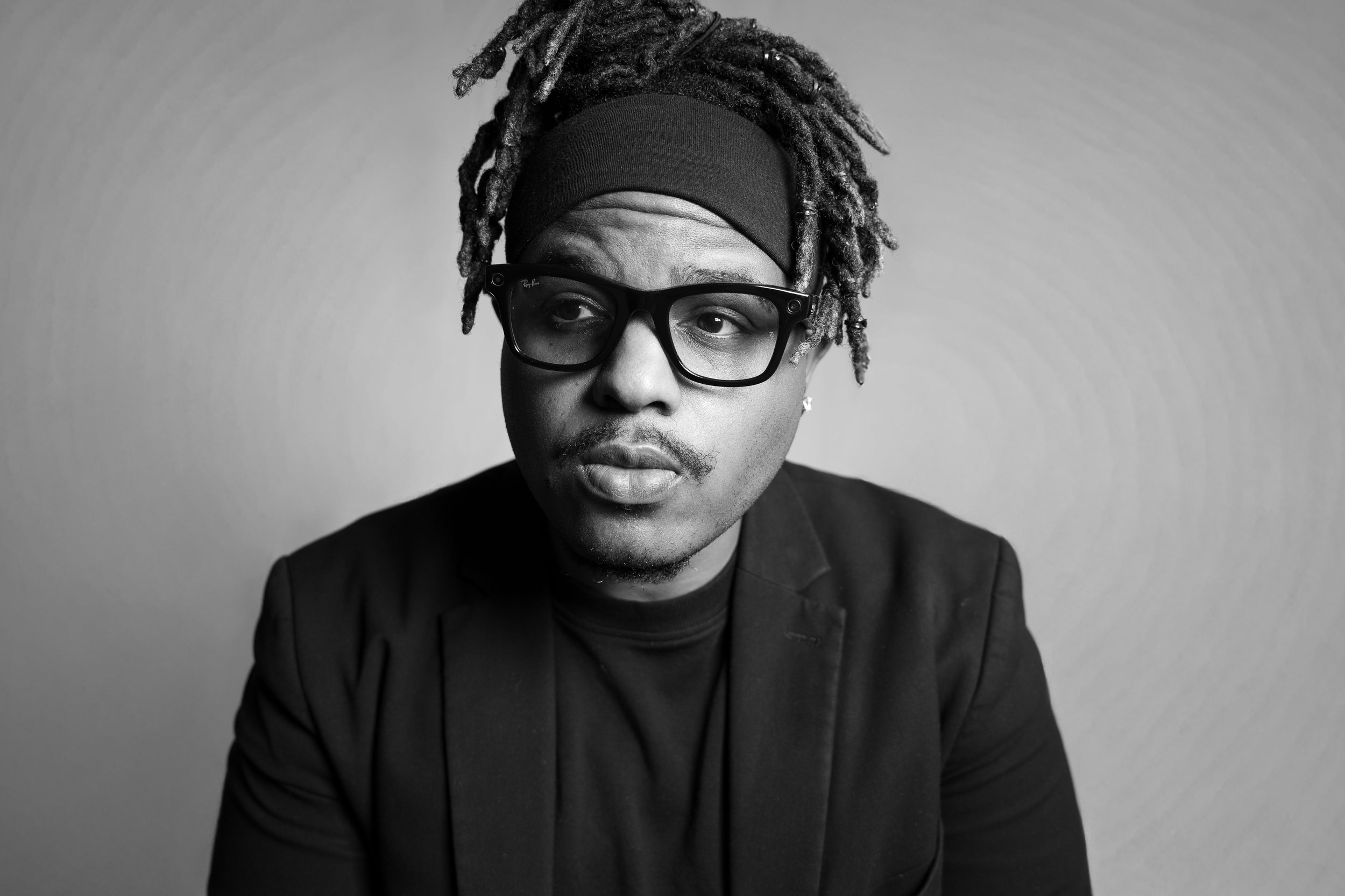 A black and white portrait of a man with dreadlocks wearing glasses, a headband, a black blazer, and a T-shirt, looking to the side.