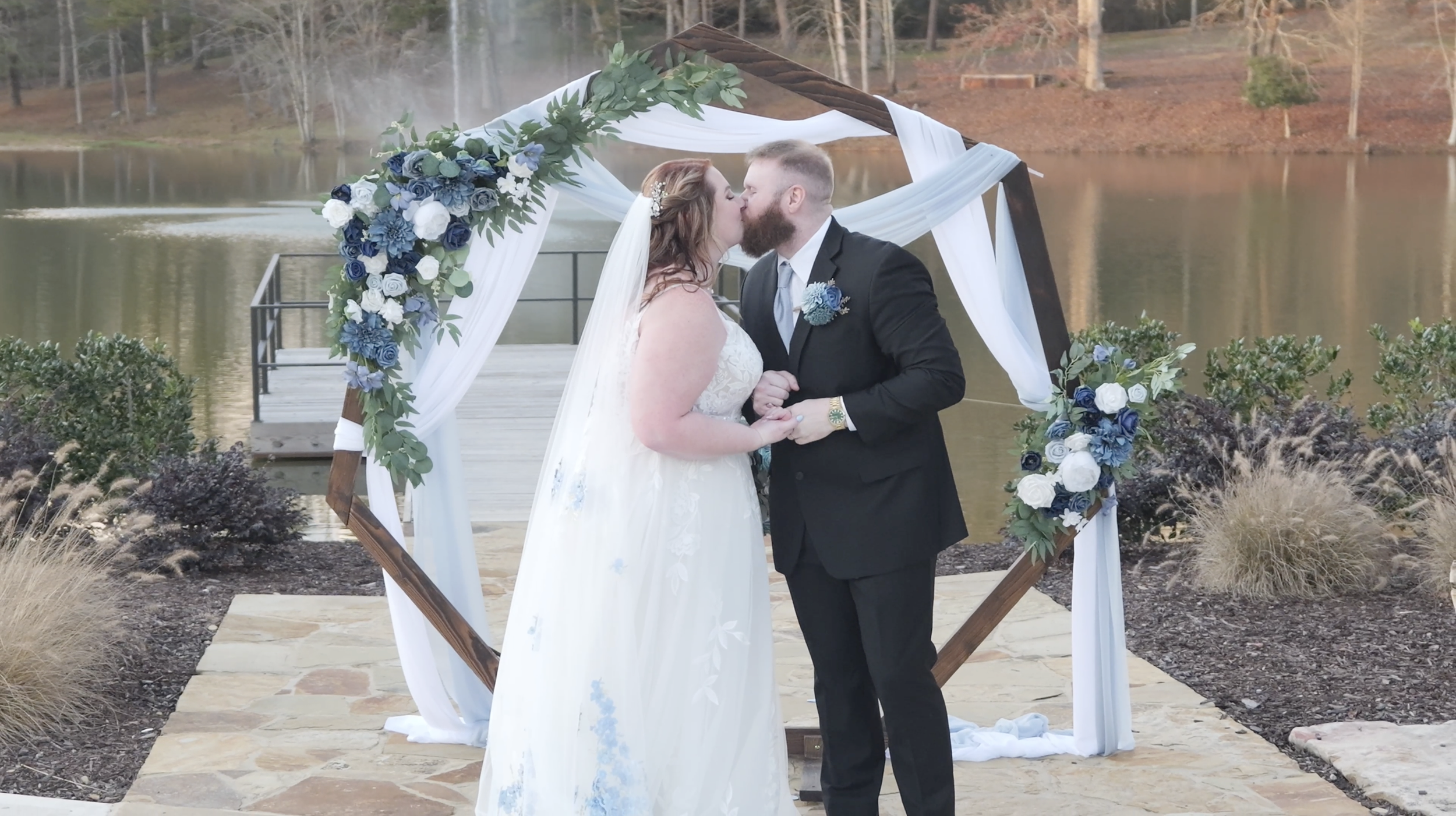 A wedding couple sharing a kiss under a decorated arch by a lake with yachts and trees in the background.