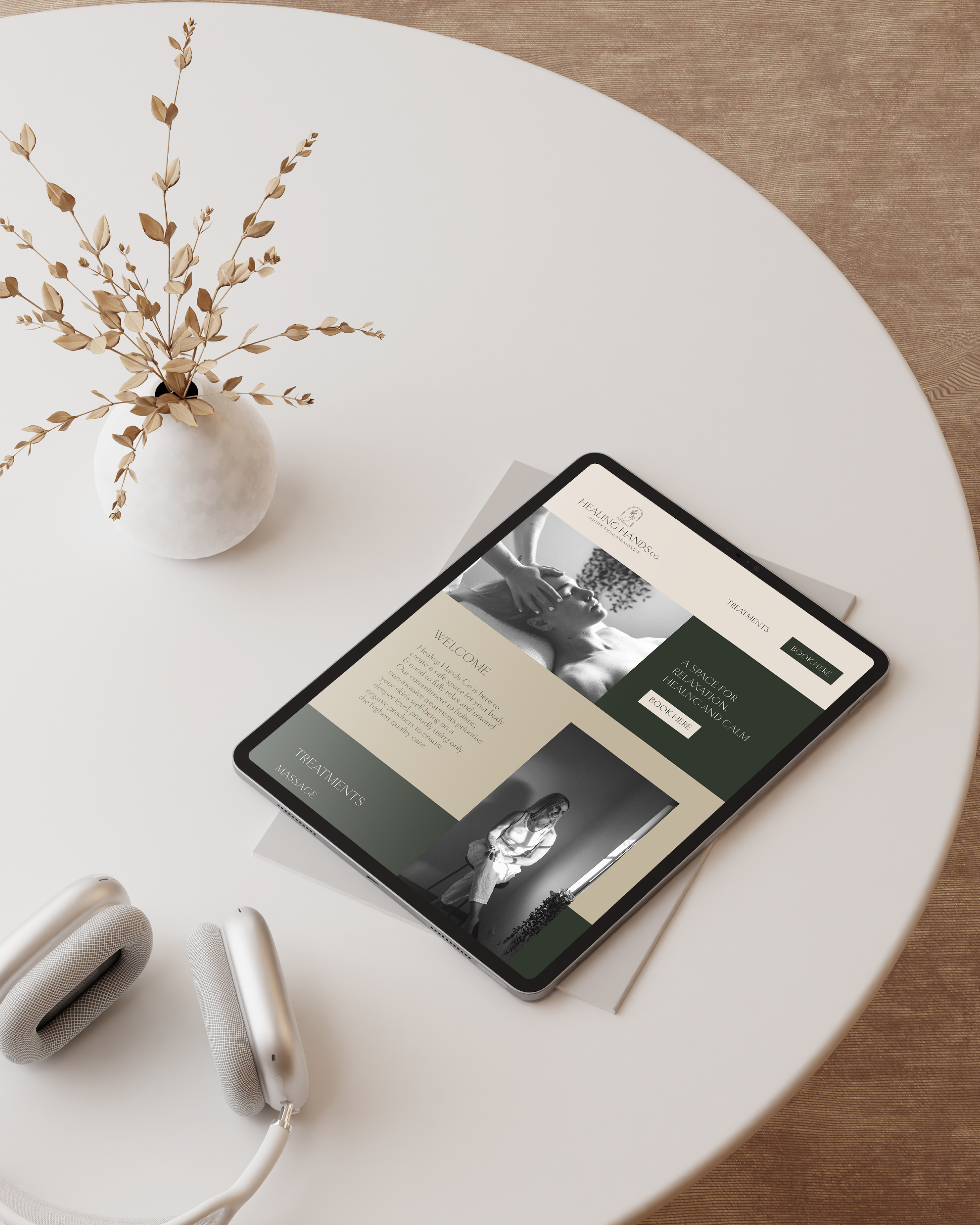 Tablet displaying a website for Healing Hands, a massage therapy service, placed on a round white table with a white vase containing dried branches, and a pair of white over-ear headphones nearby.