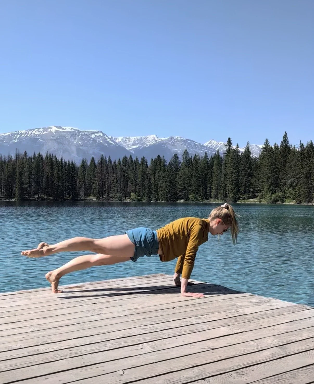 Image of a female doing Pilates movements on a dock by a turquoise blue lake with trees and mountains in the background.