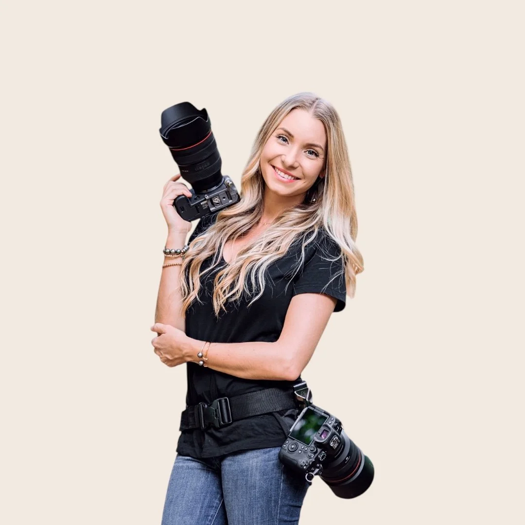 A smiling woman with long blonde hair holding a professional camera with a large telephoto lens, wearing a pink dress and bracelets, against a plain light background.