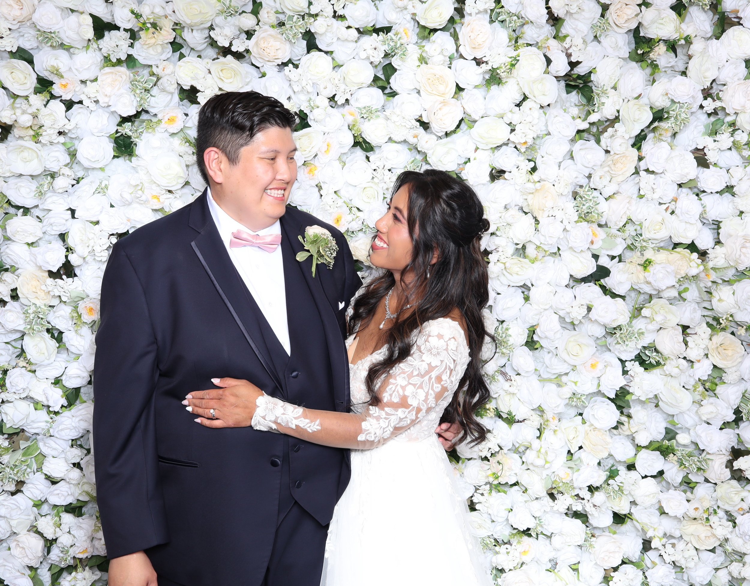 A newlywed couple dressed in wedding attire standing in front of a wall covered with white roses. The groom is wearing a tuxedo with a light pink bow tie and boutonniere. The bride is in a white lace wedding gown with long sleeves and a necklace. They are smiling at each other lovingly.