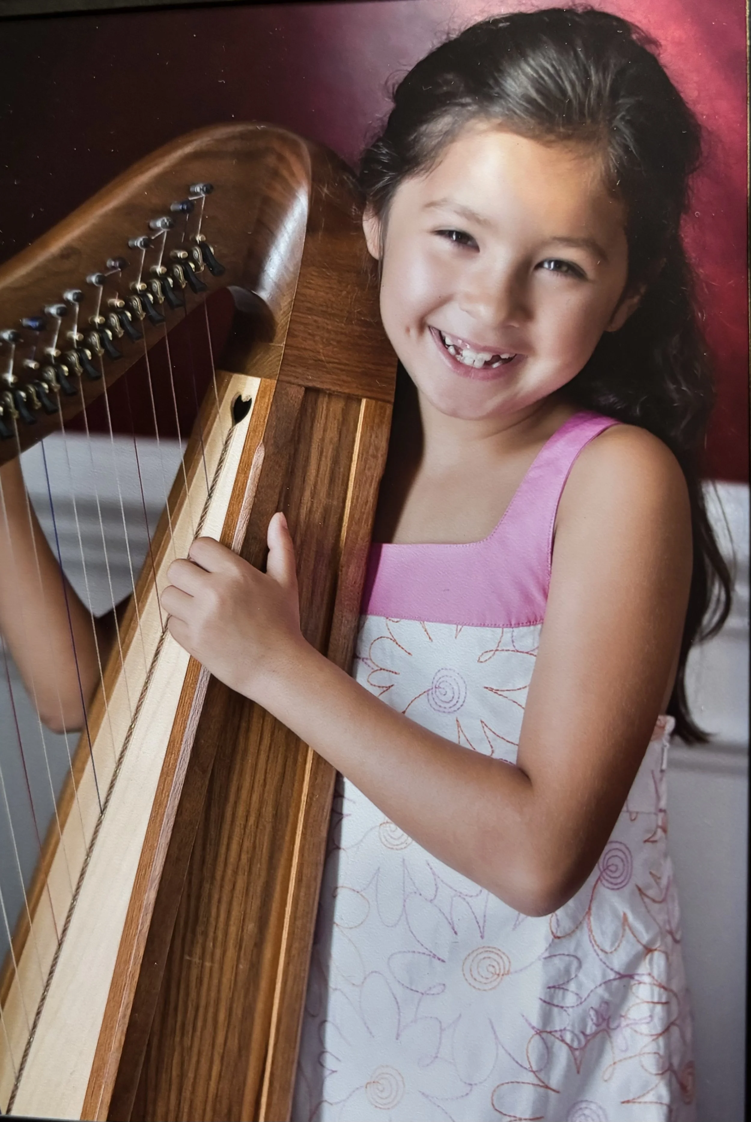 A young girl with dark hair, wearing a pink and white dress, smiling and holding a wooden harp.