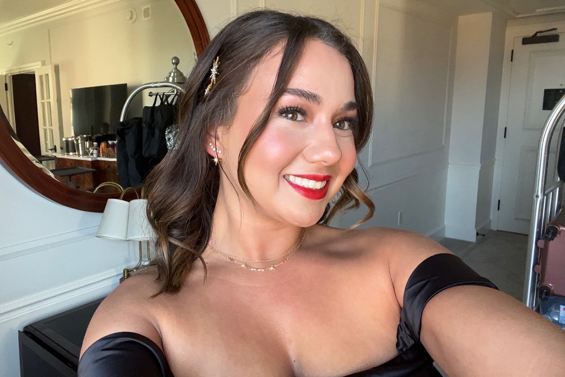 A smiling woman with shoulder-length brown hair styled in loose waves, wearing a black off-the-shoulder top, red lipstick, and gold jewelry, taking a selfie in a well-lit room.