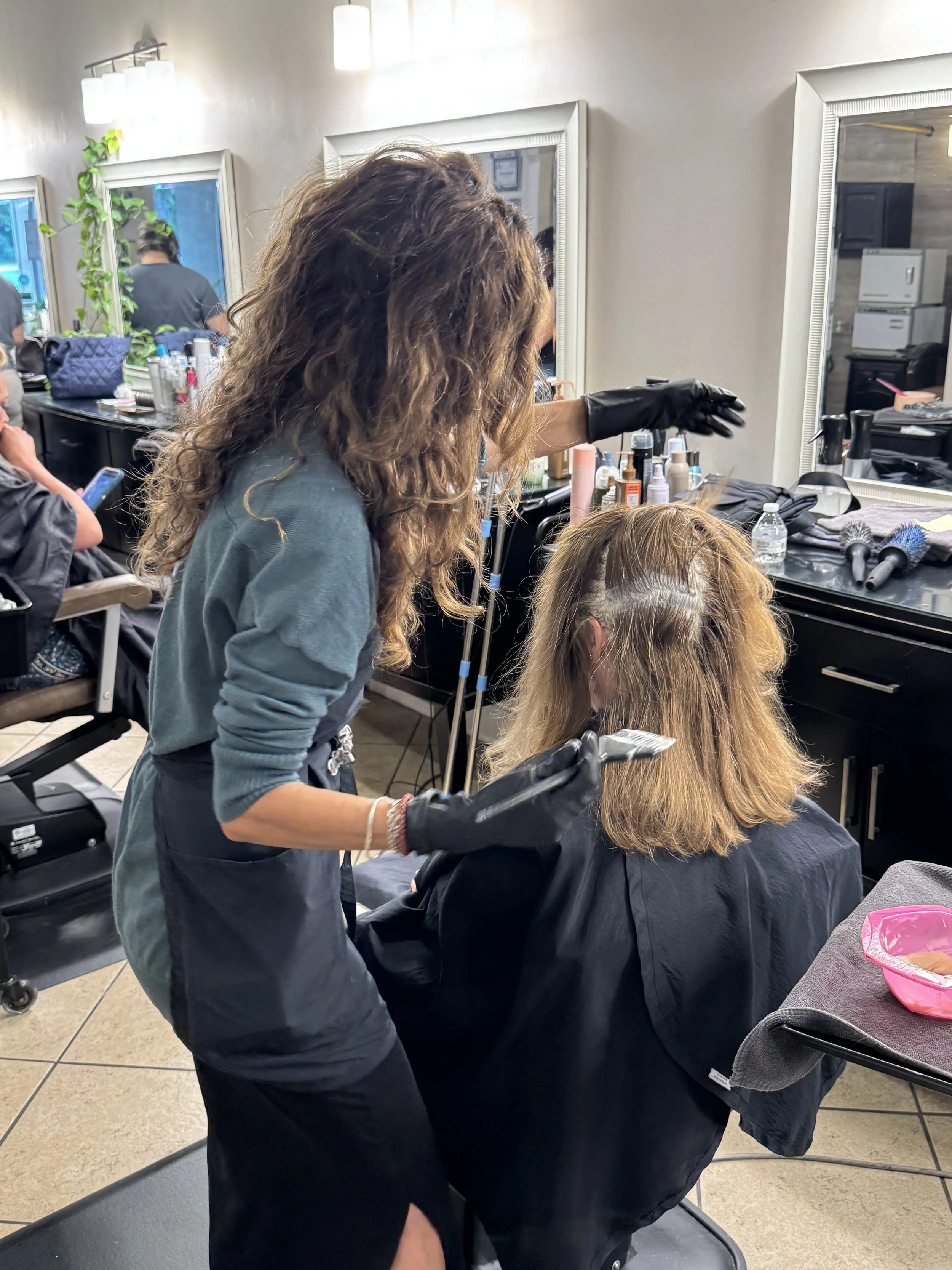 A hairstylist with curly hair and black gloves is styling a woman's hair in a salon, with mirrors, hair products, and styling tools visible in the background.