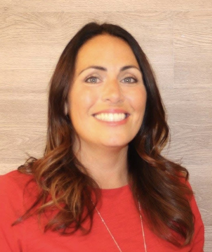 Smile woman with long brown hair in a red shirt standing in front of a wooden textured wall.