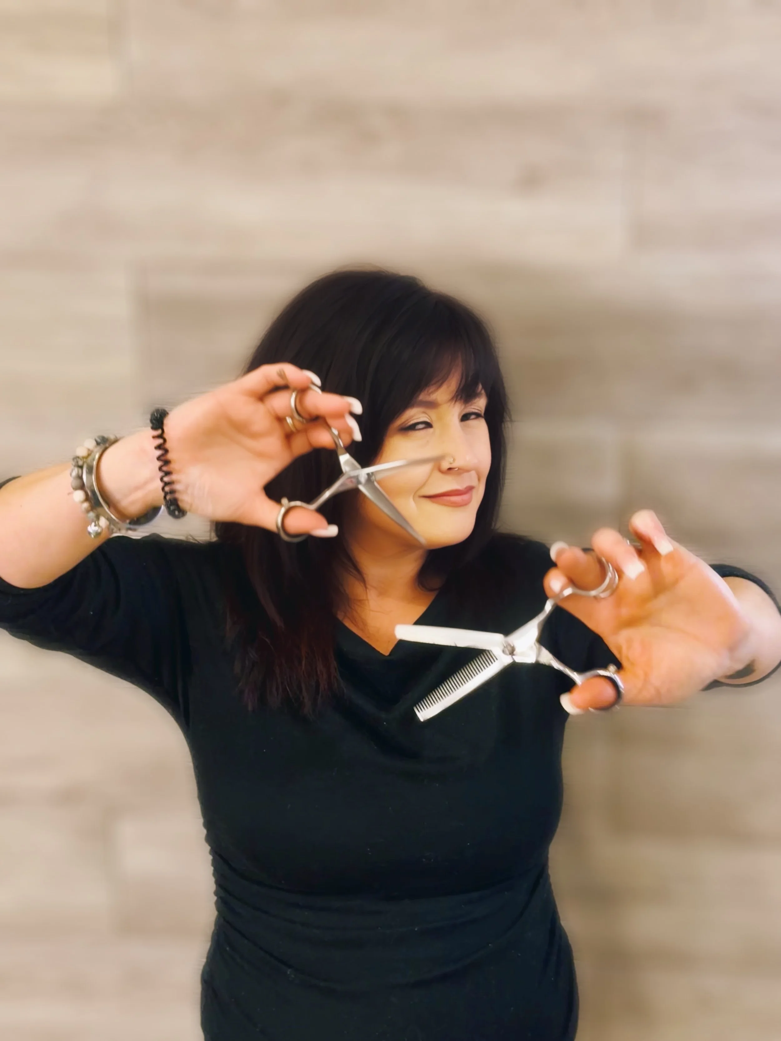 A woman with dark hair and multiple bracelets on her wrist, holding a pair of scissors and a comb, smiling playfully toward the camera.