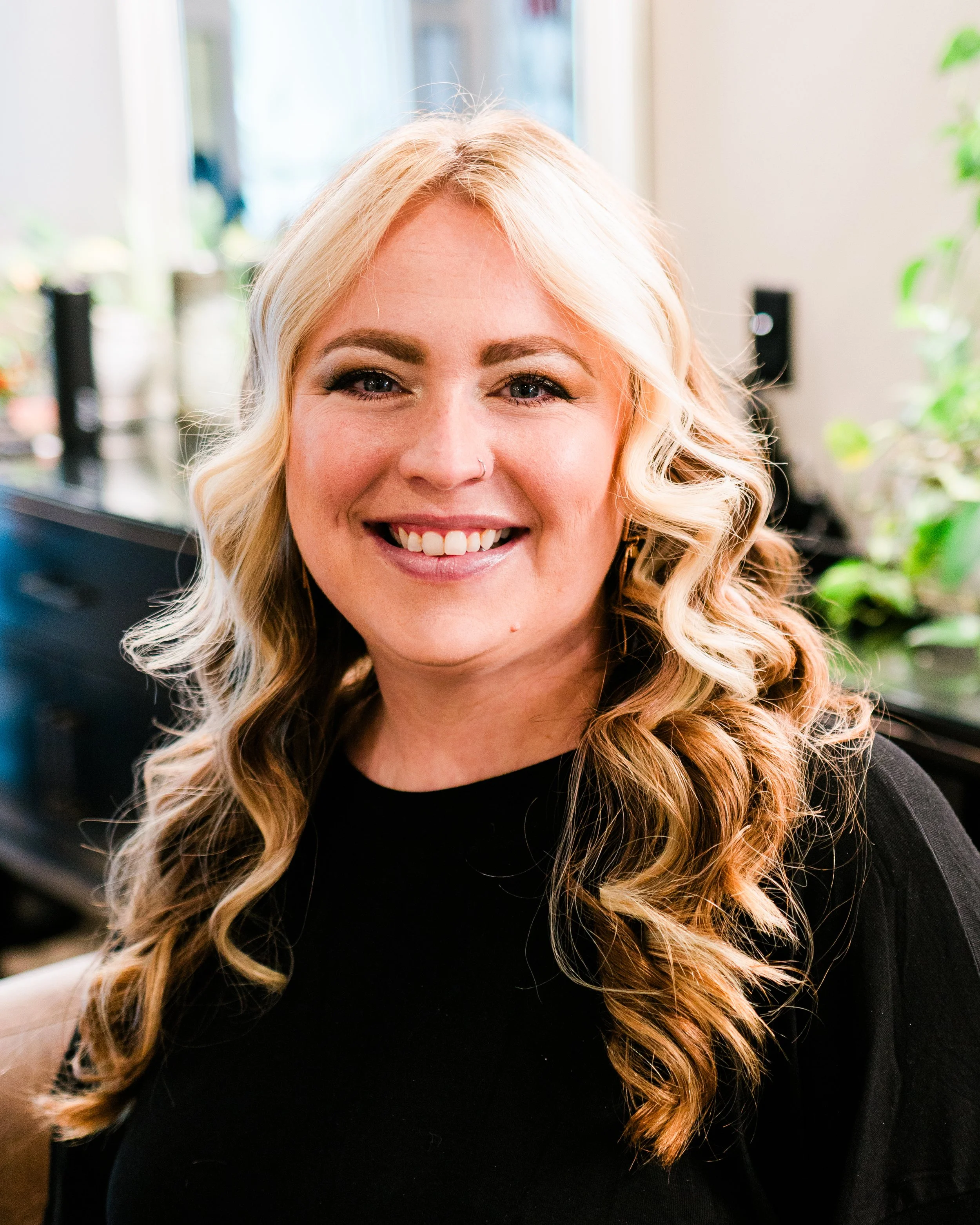 A woman with long blonde curly hair smiling indoors, wearing a black top.