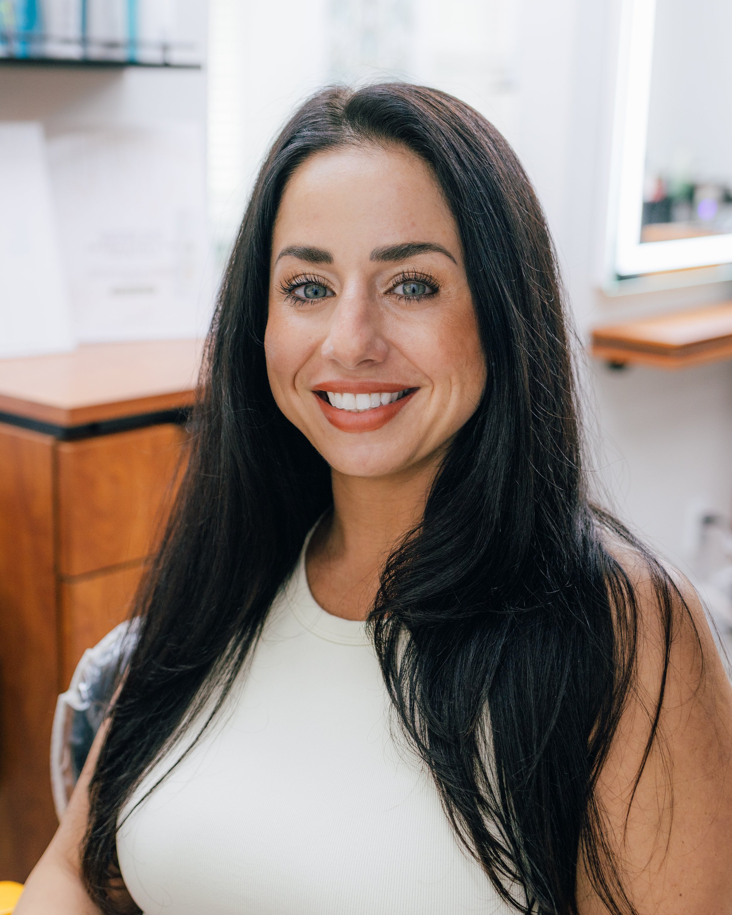 A woman with long black hair and blue eyes smiling in an office setting.