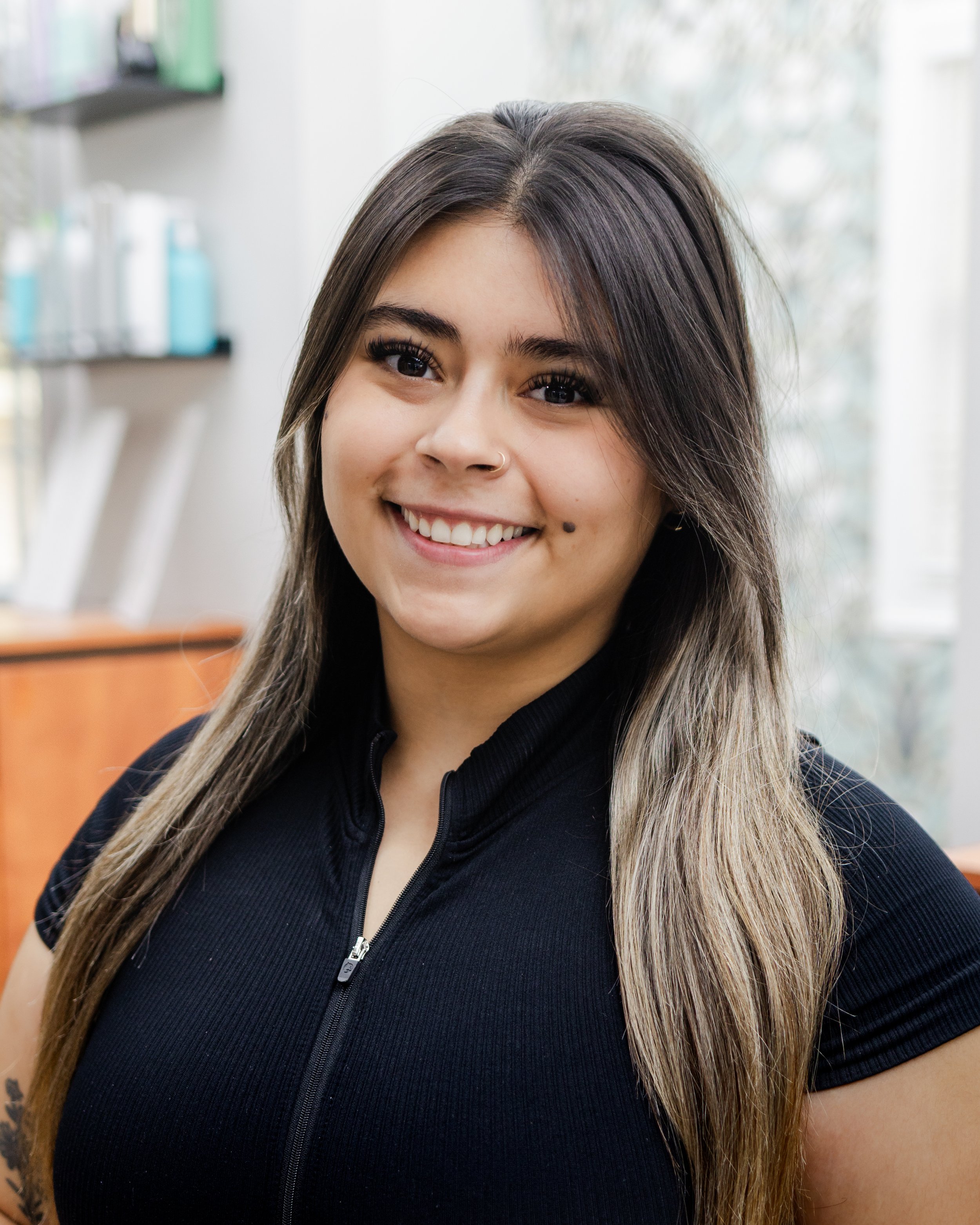 Young woman with long hair, smiling in a bright indoor setting with shelves of products in the background.