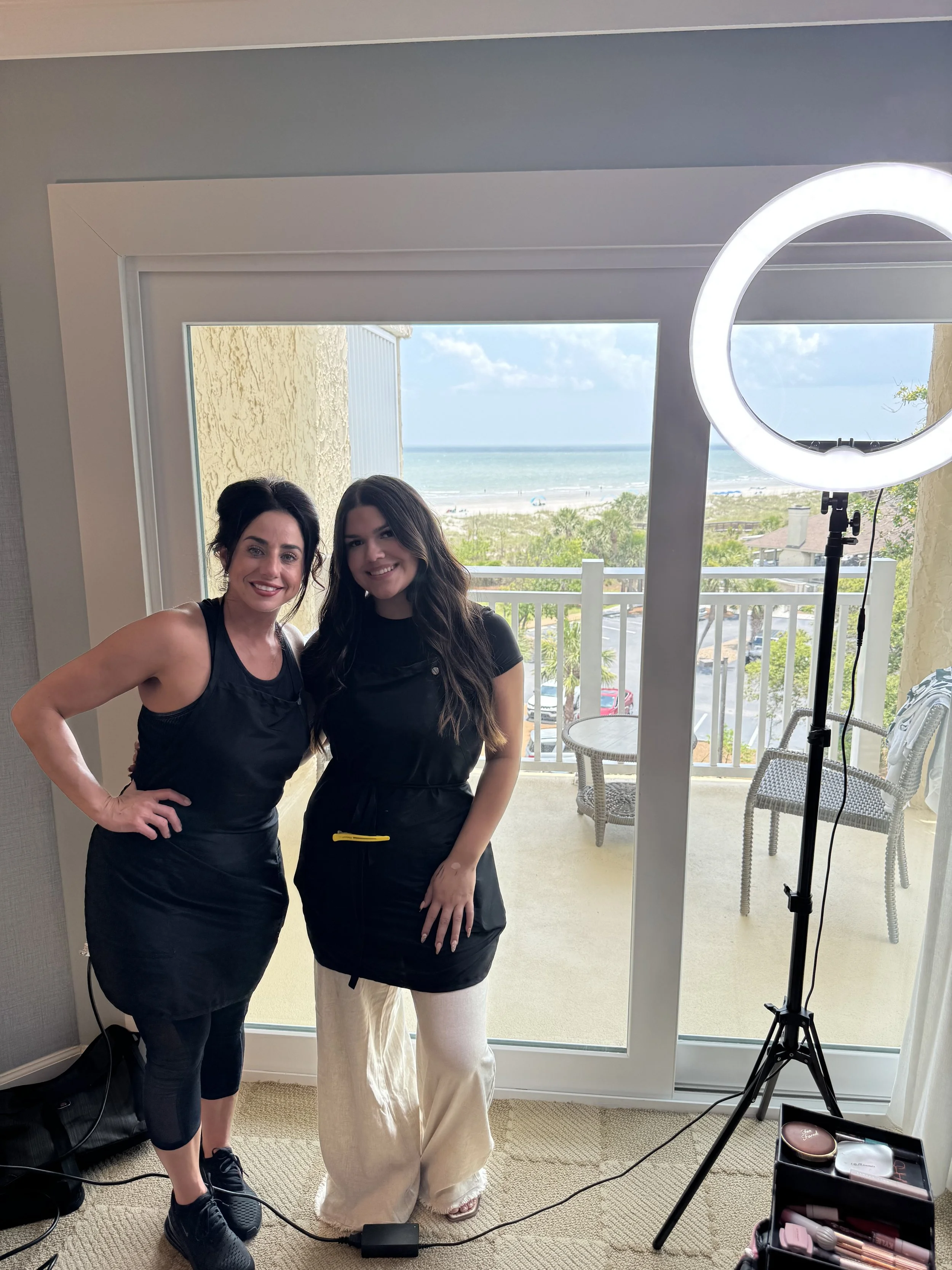 Two women standing in front of a large window overlooking a beach, with a ring light and makeup supplies nearby.