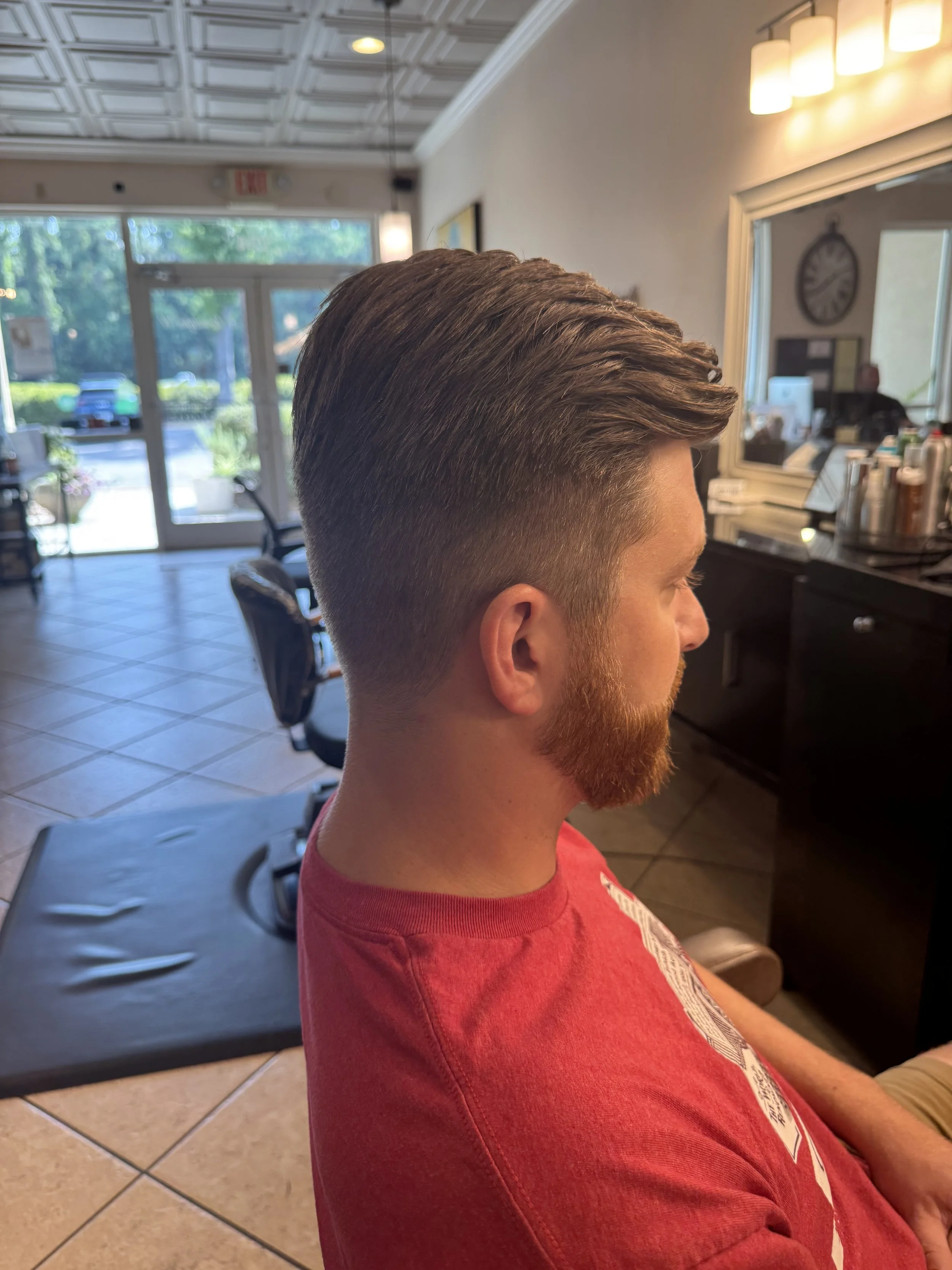 A young man with a thick, styled haircut and a reddish beard sitting in a barber shop, facing sideways. The background shows a door, large windows, a clock, and a mirror reflecting the interior of the shop.