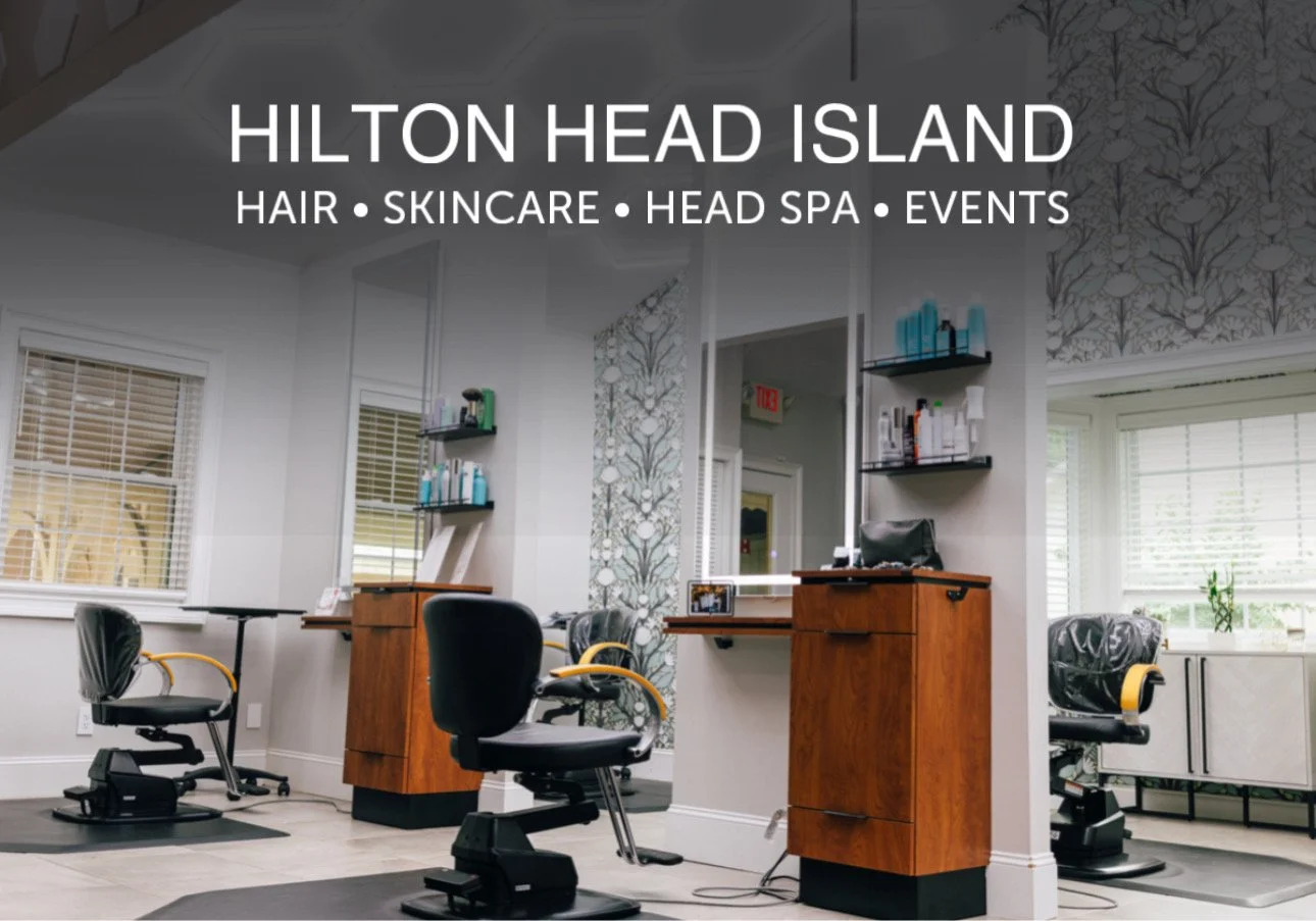 Interior view of a hair salon with three styling chairs, mirrors, and shelves with hair products. Natural light from large windows with blinds. Wooden furniture and decorative wallpaper.