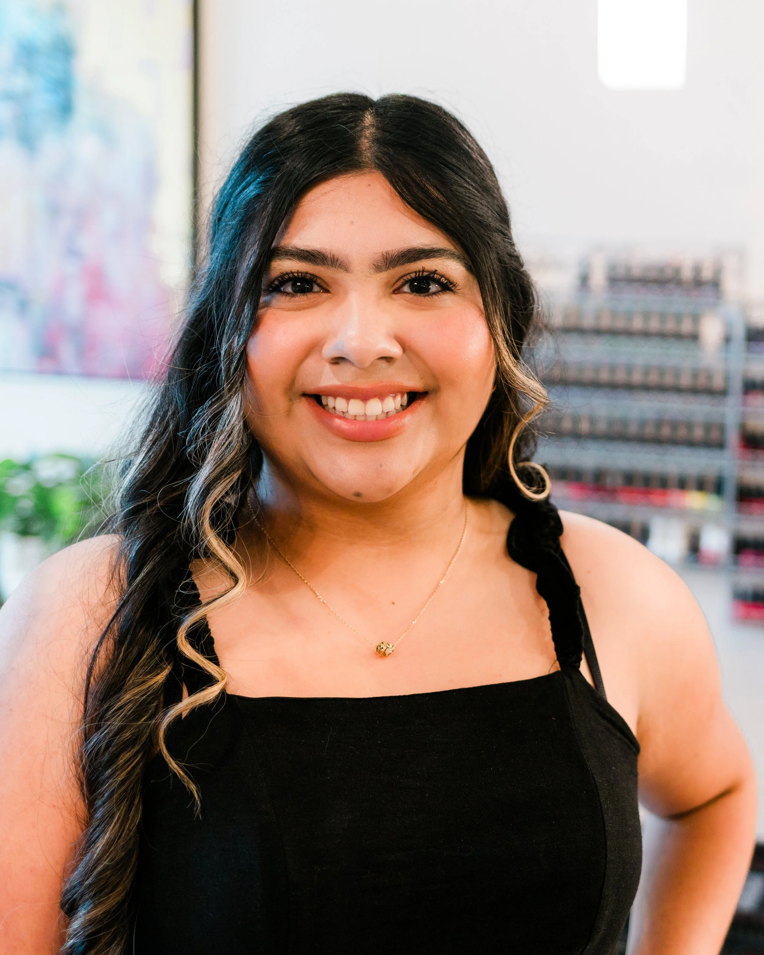 A young woman with long, dark hair and a bright smile, wearing a black top with ruffled straps, standing indoors with a cityscape in the background.