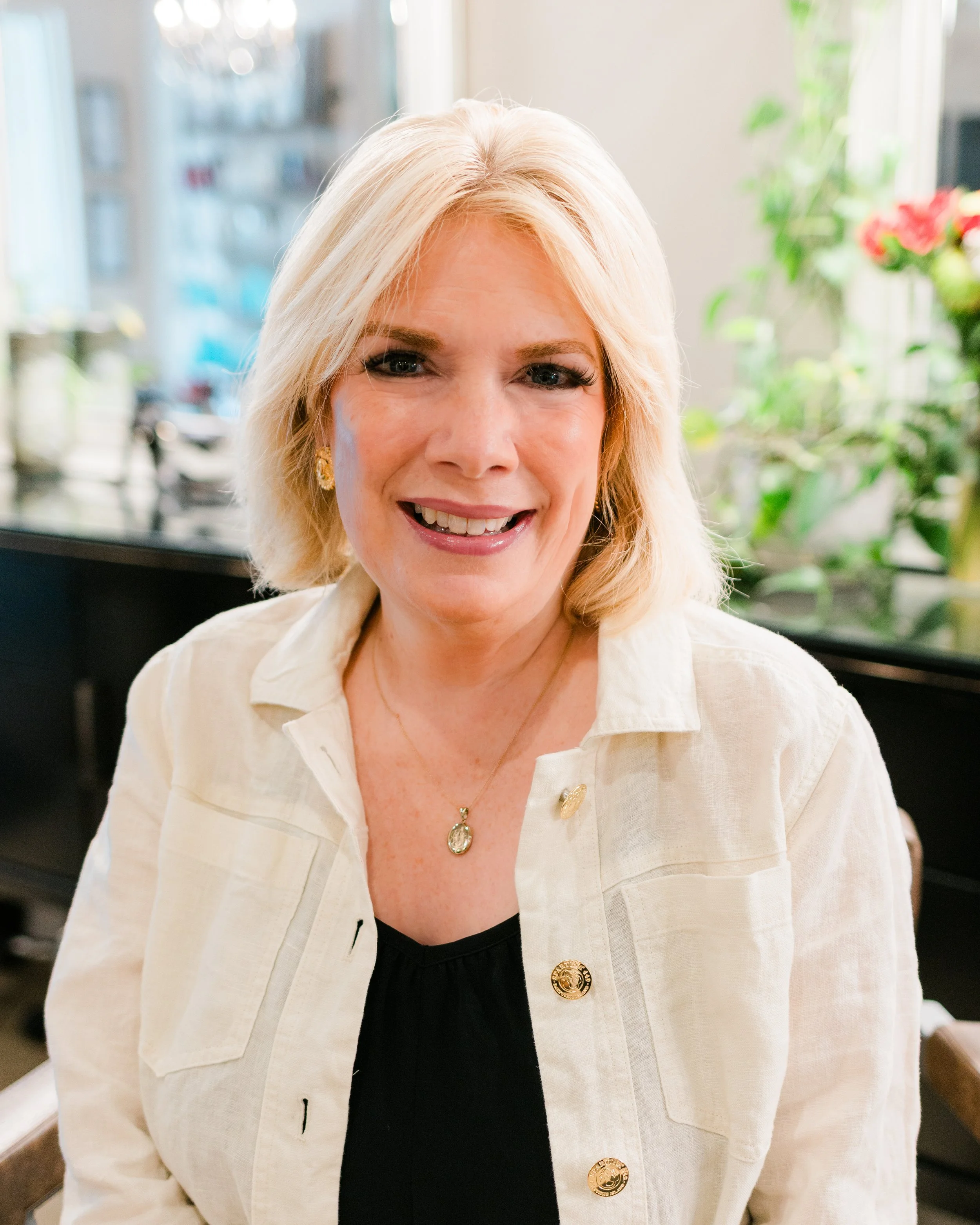 A woman with blonde hair smiling, wearing a cream-colored jacket with gold buttons and jewelry, sitting indoors near a window with plants and flowers in the background.