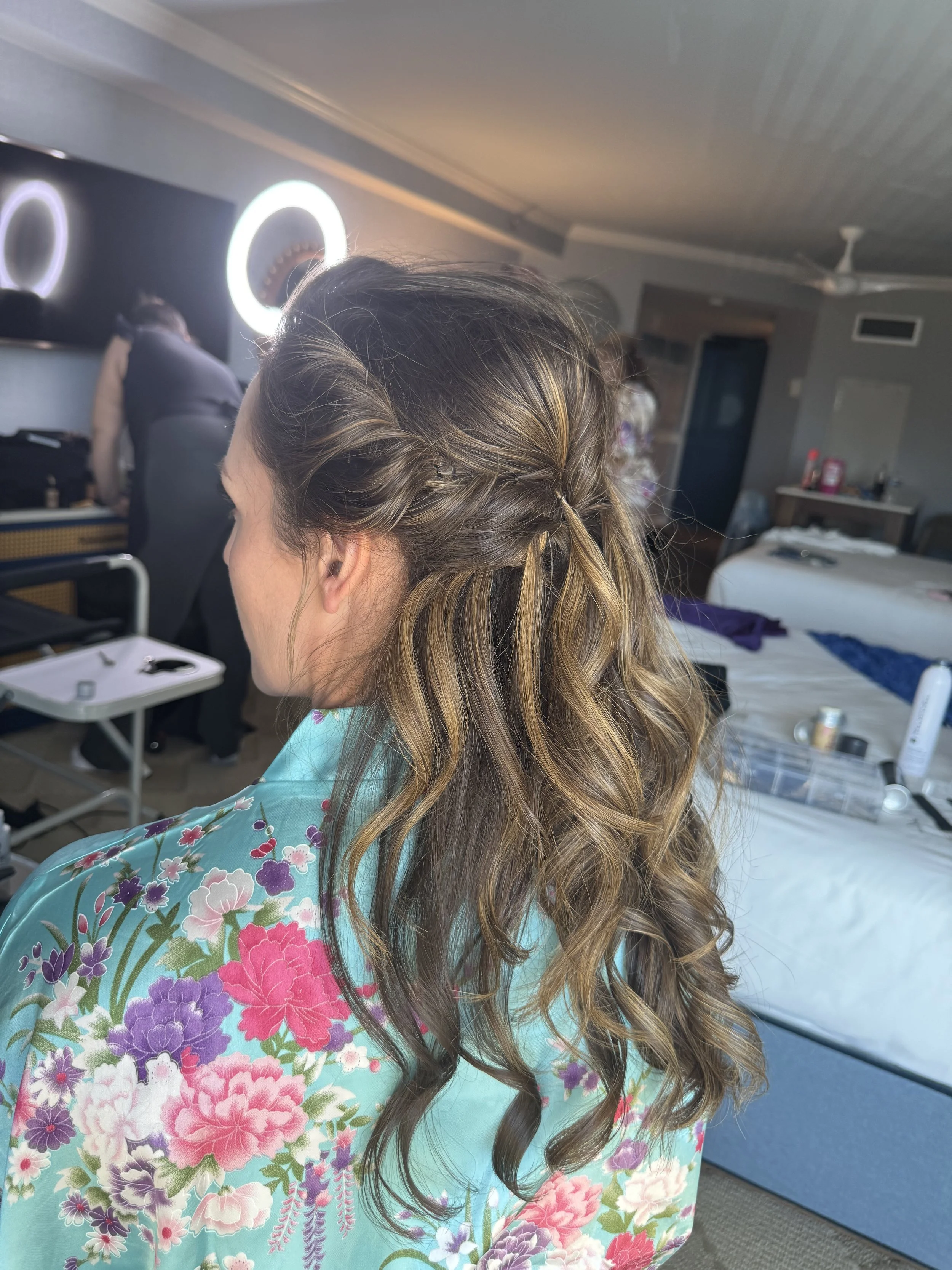 Back view of a woman with long, curly hair featuring blonde highlights, wearing a teal floral kimono, in a room with a bed and people in the background.