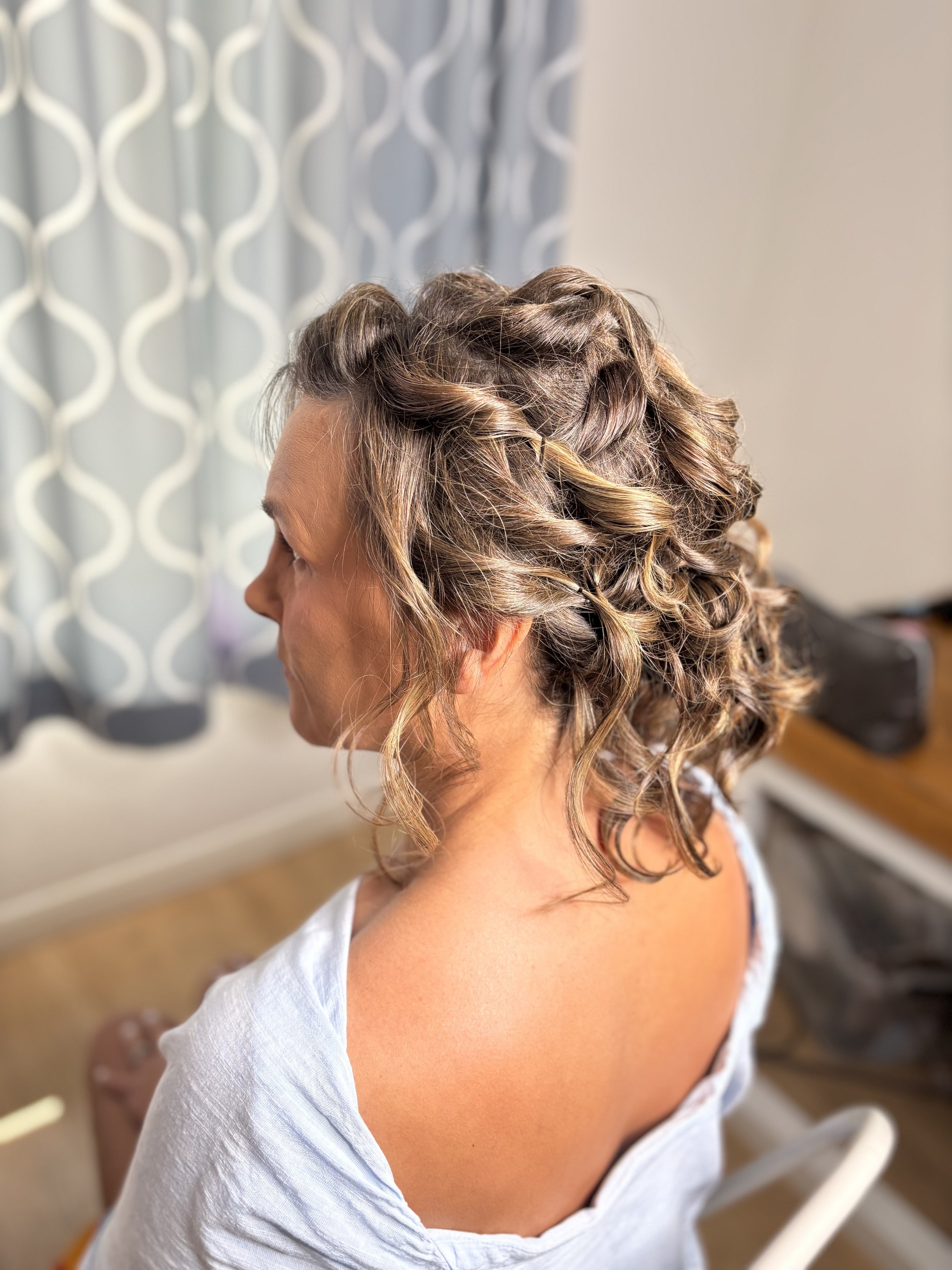 A woman with shoulder-length, curly hair styled with twists and highlights, sitting indoors near a window with patterned curtains.