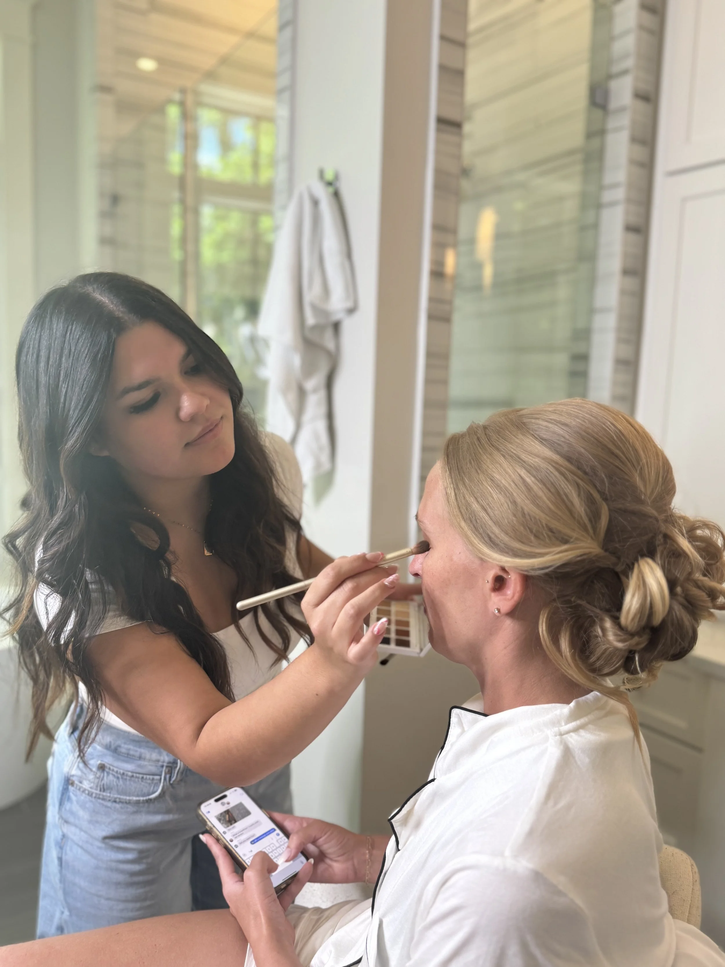 A woman with blonde hair is sitting while another woman applies makeup around her eyes with a brush. The woman receiving makeup is holding a phone with a messaging app open. They are in a well-lit room with wood-paneled walls and a towel hanging in the background.