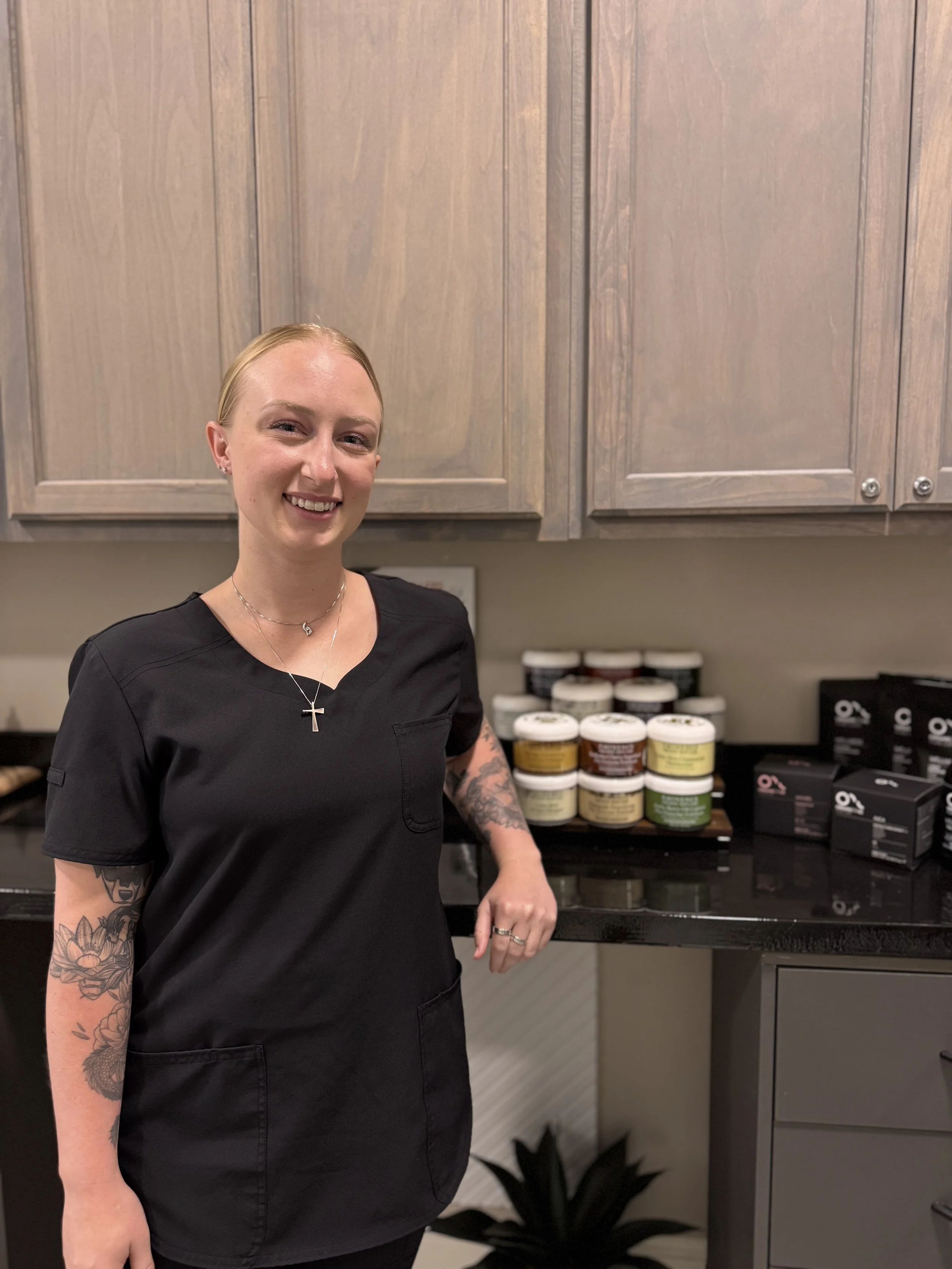 A woman with blonde hair and tattoos wearing a black medical scrub top, smiling, standing in front of a countertop with various jars and boxes of health or beauty products, in a room with wood cabinets.