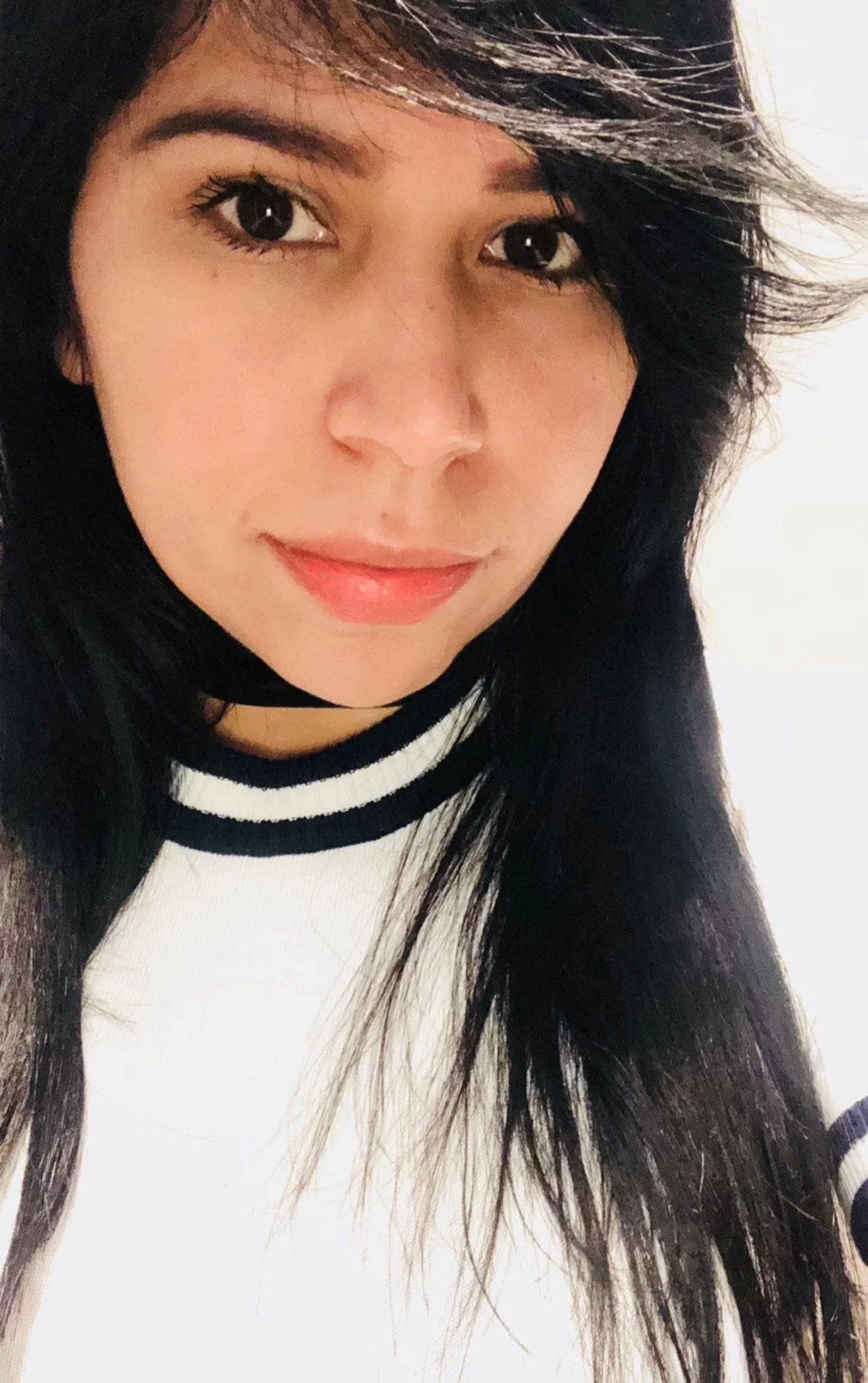 Close-up of a young woman with long dark hair, smiling slightly, wearing a white top with dark stripes around the collar.