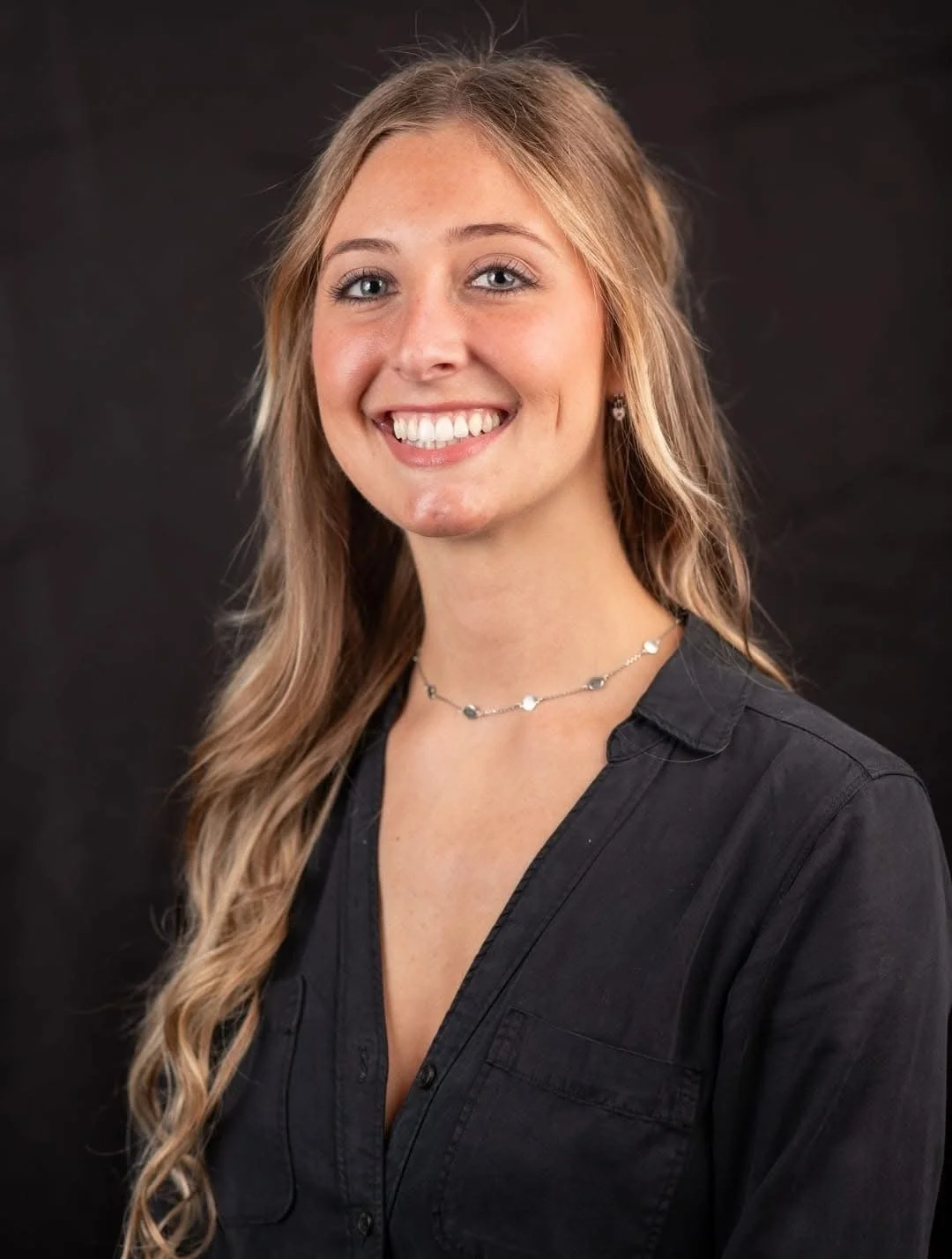 A young woman with long, wavy blonde hair, blue eyes, and fair skin, smiling, wearing a black collared shirt and a silver necklace, against a dark background.
