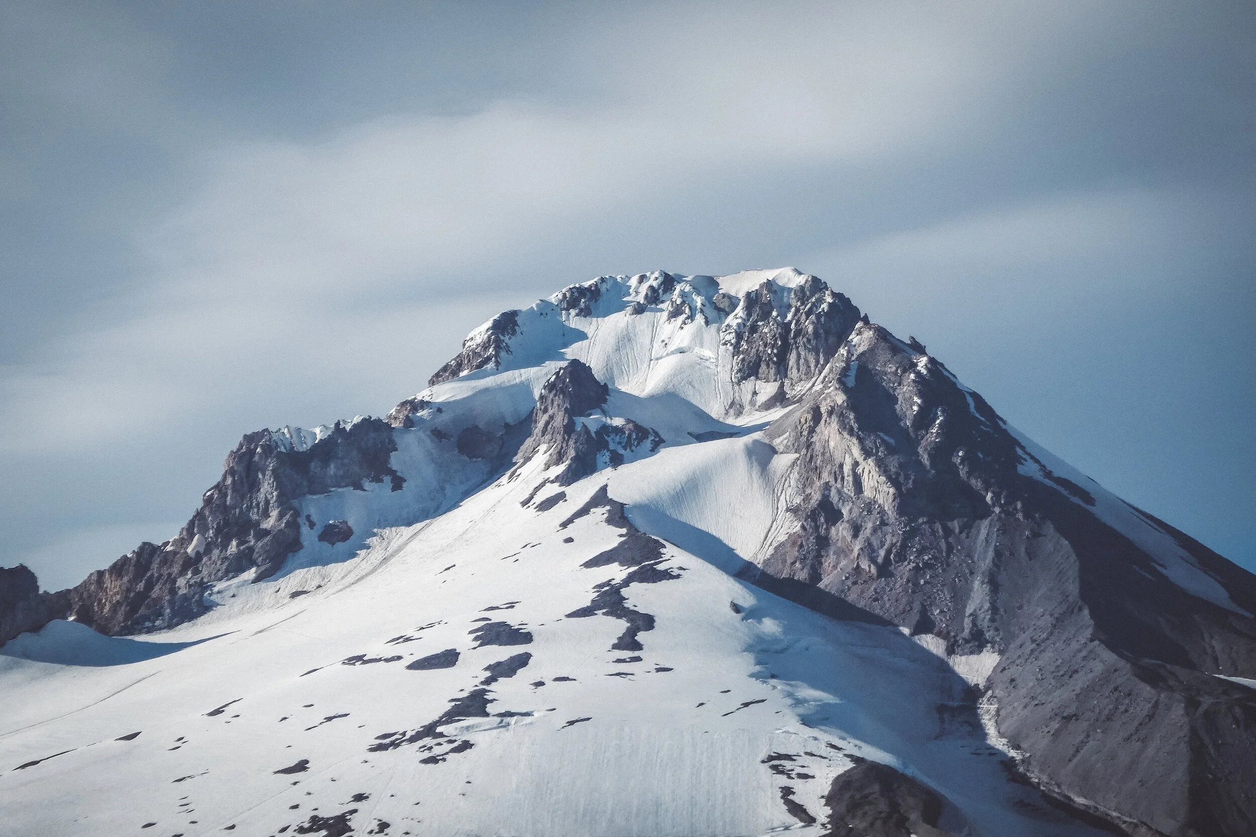 Snow-covered mountain peak with rocky ridges and a cloudy sky.