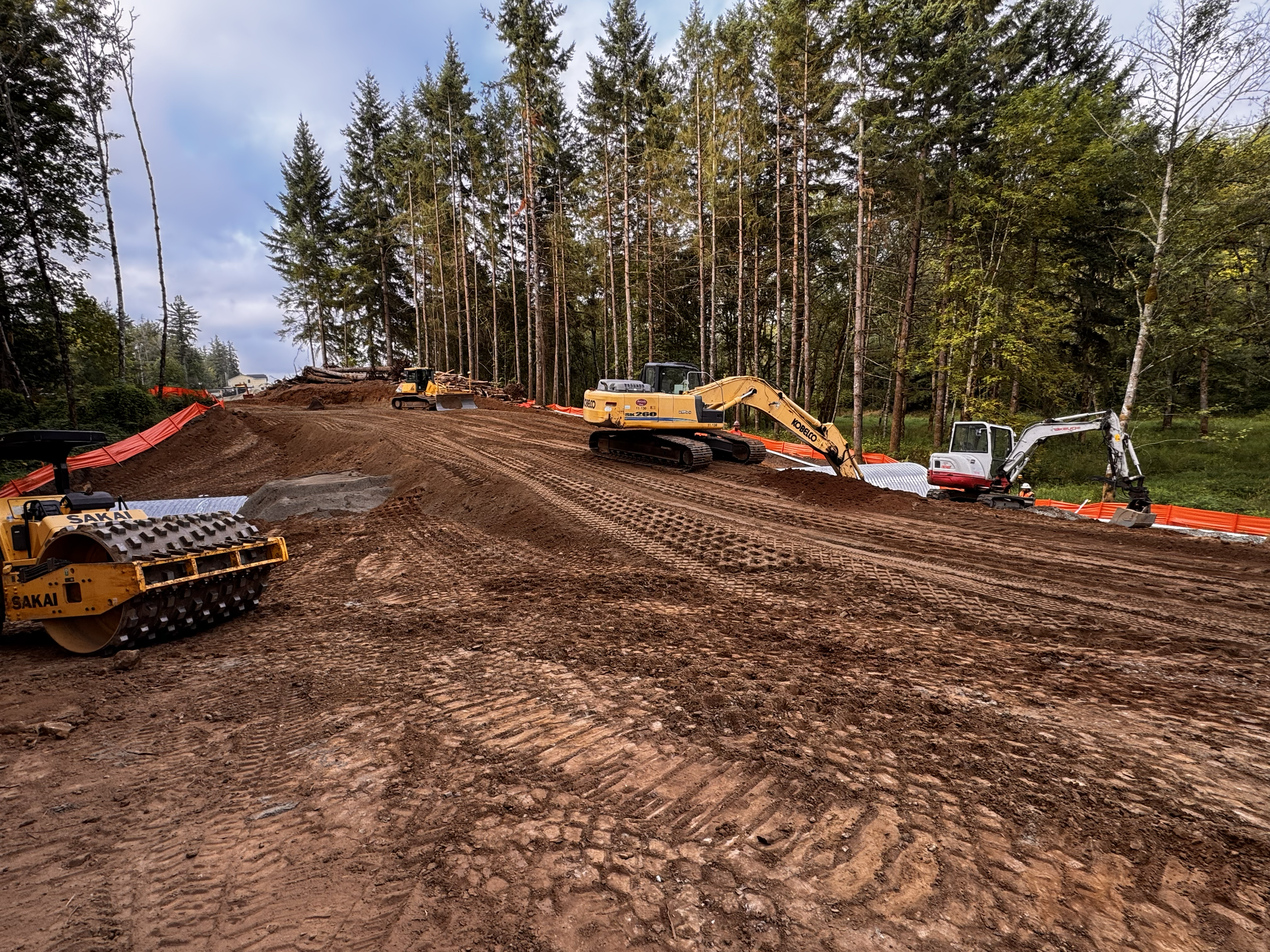 Construction site in a forest with dirt, earth-moving machinery, and orange safety barriers.