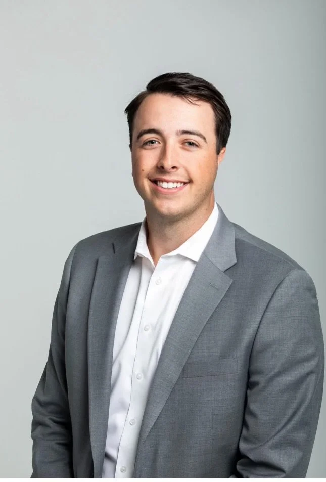 Young man in a gray suit and white shirt smiling against a plain background.