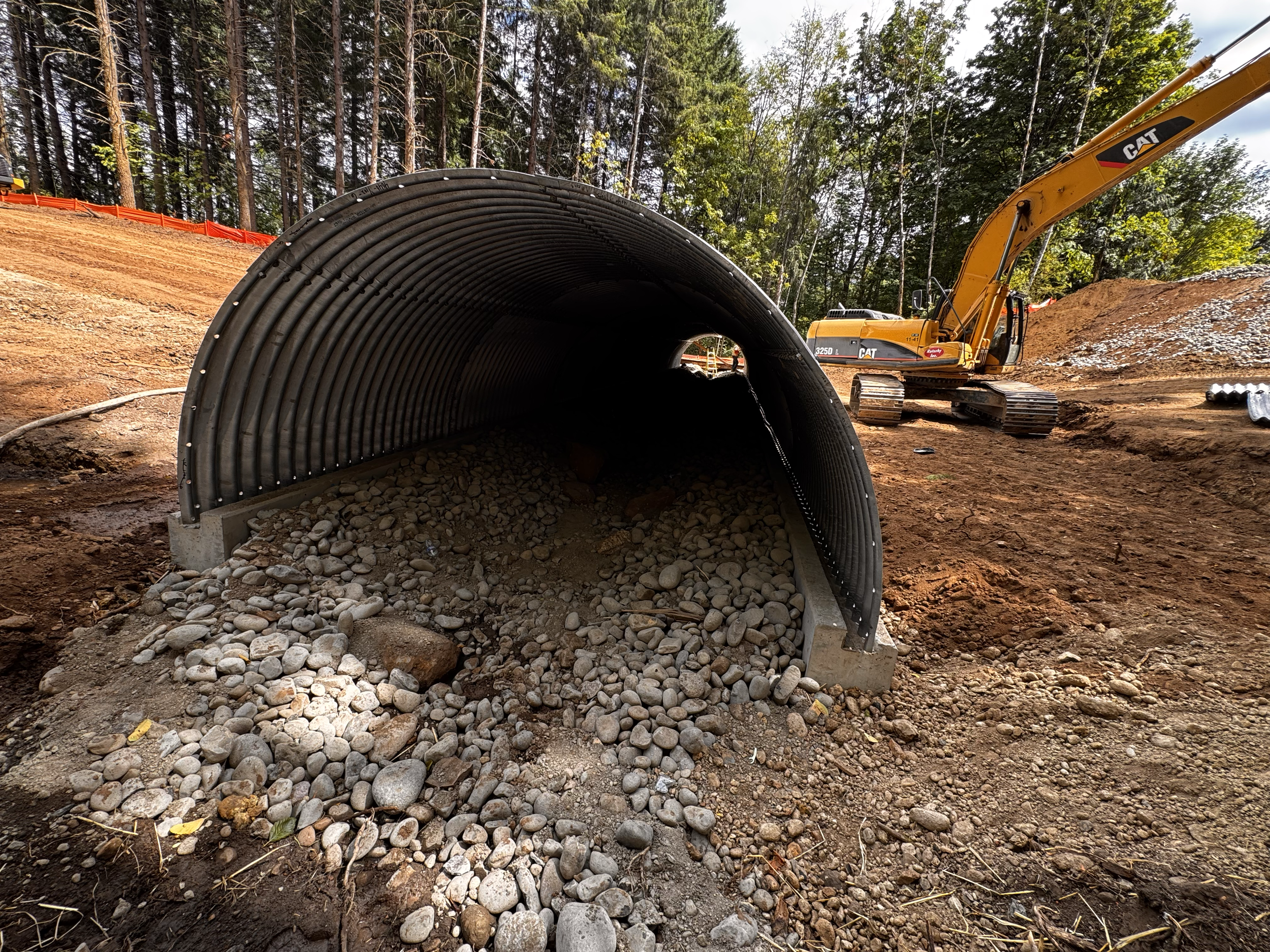 A construction site with a large corrugated pipe and an excavator in a wooded area.