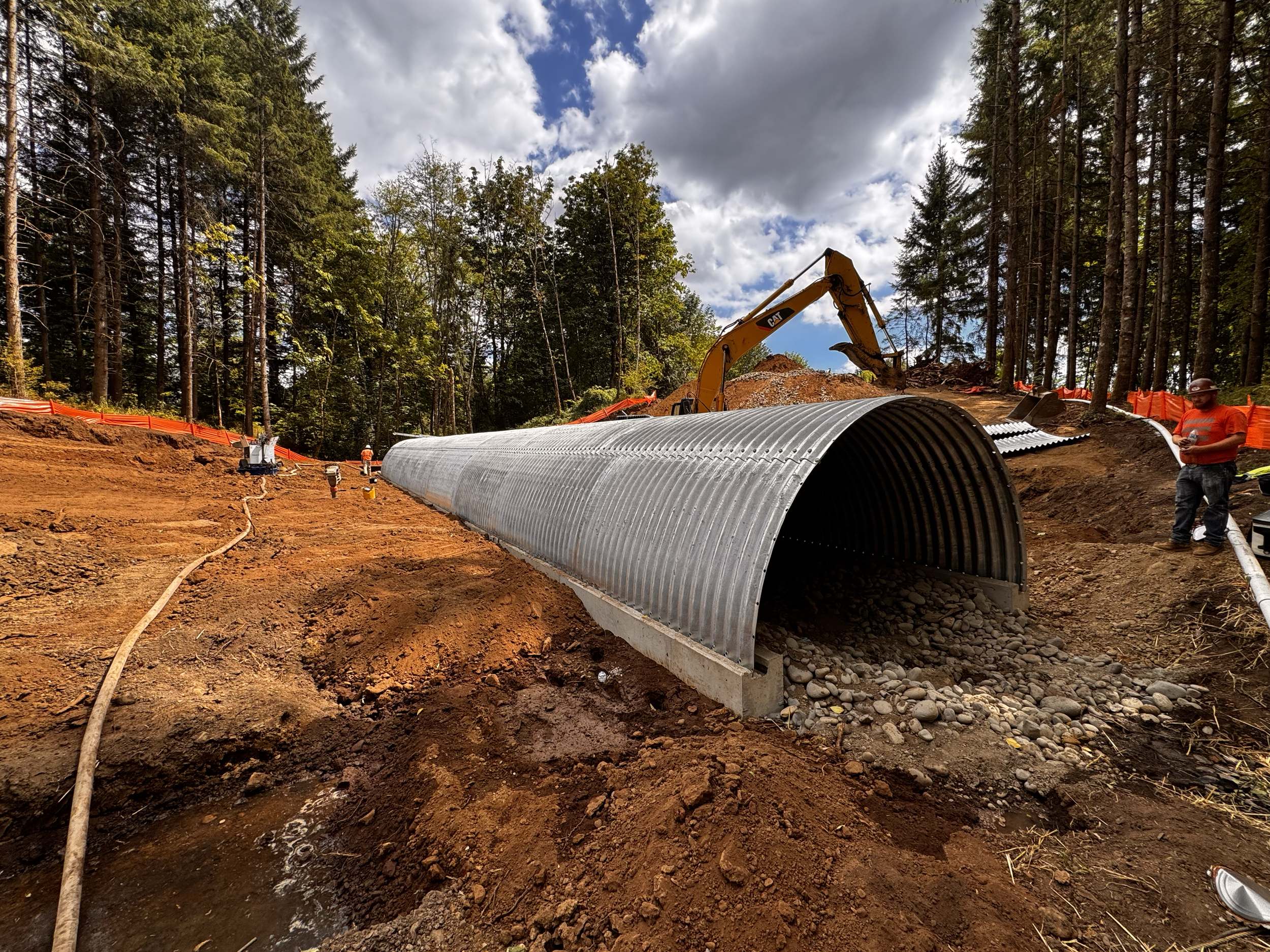 Construction site in a forest with a large corrugated metal pipe for drainage, excavator, and workers installing infrastructure.