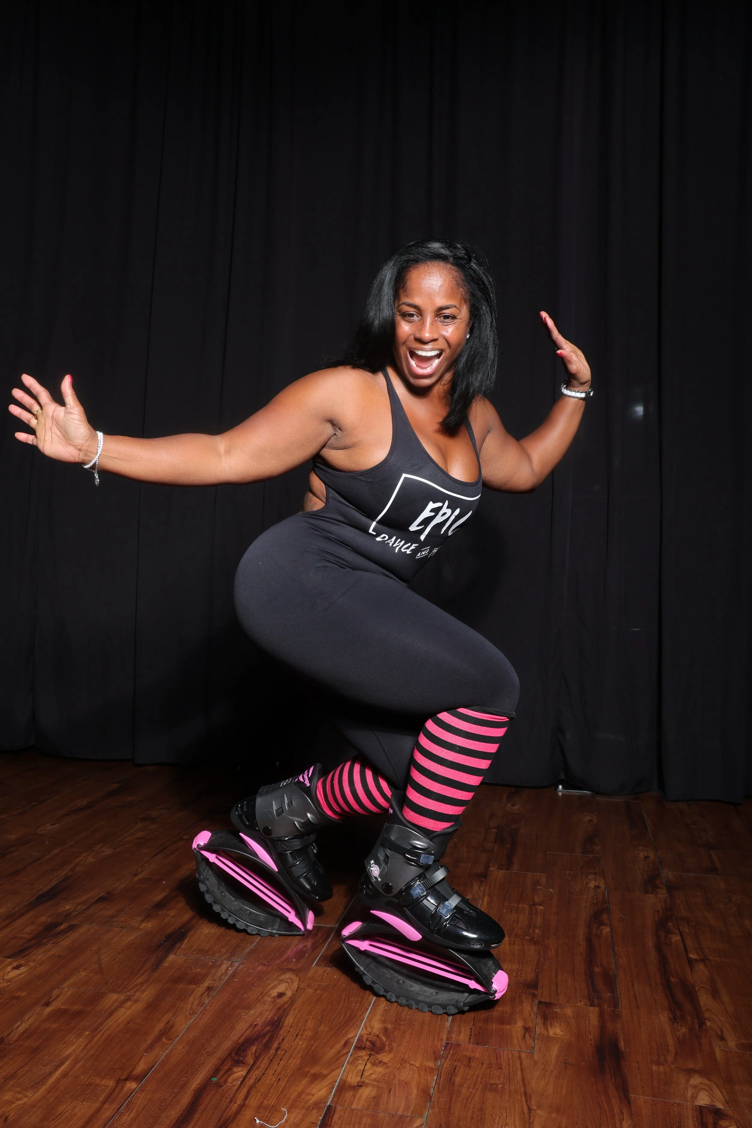 A woman in black rollerblades, wearing striped pink and black socks, black workout clothing, smiling, and posing in front of black curtains on a wooden floor.