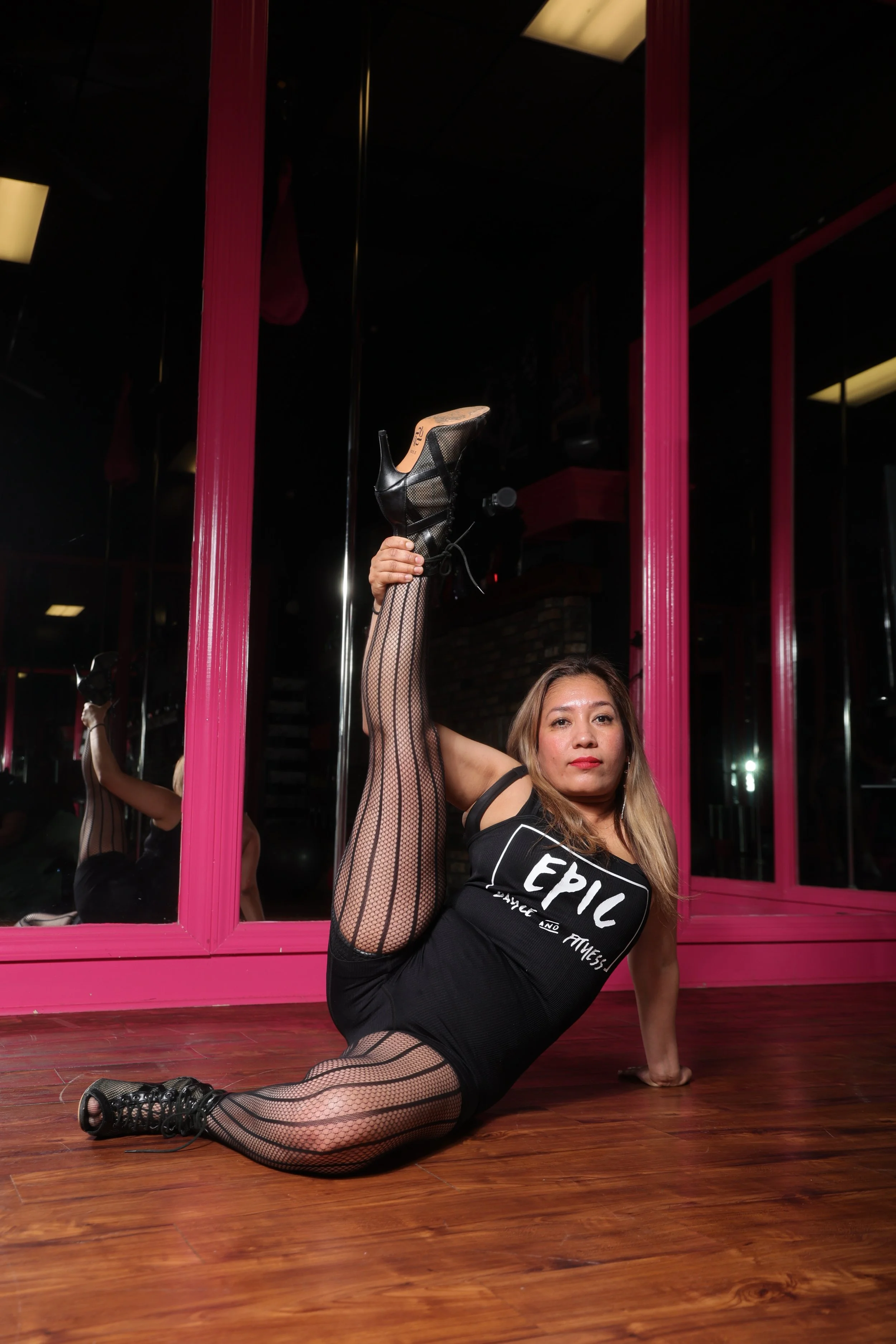 A woman posing in a dance or fitness studio, raising her leg while sitting on the floor, wearing black fishnet tights, a black shirt with white text, and black shoes, with a pink wall and mirrors reflecting her pose in the background.
