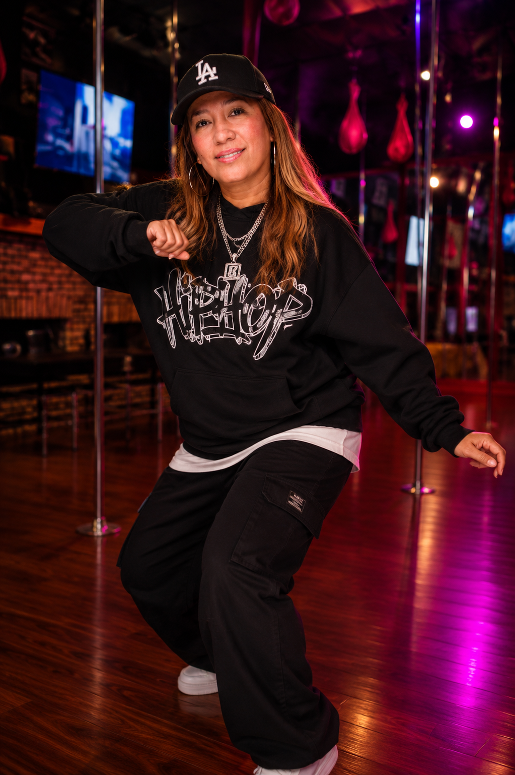 A woman in casual streetwear, including a black hoodie, black pants, and a black baseball cap, is dancing inside a dimly lit dance studio with pink lighting and stripping poles in the background.