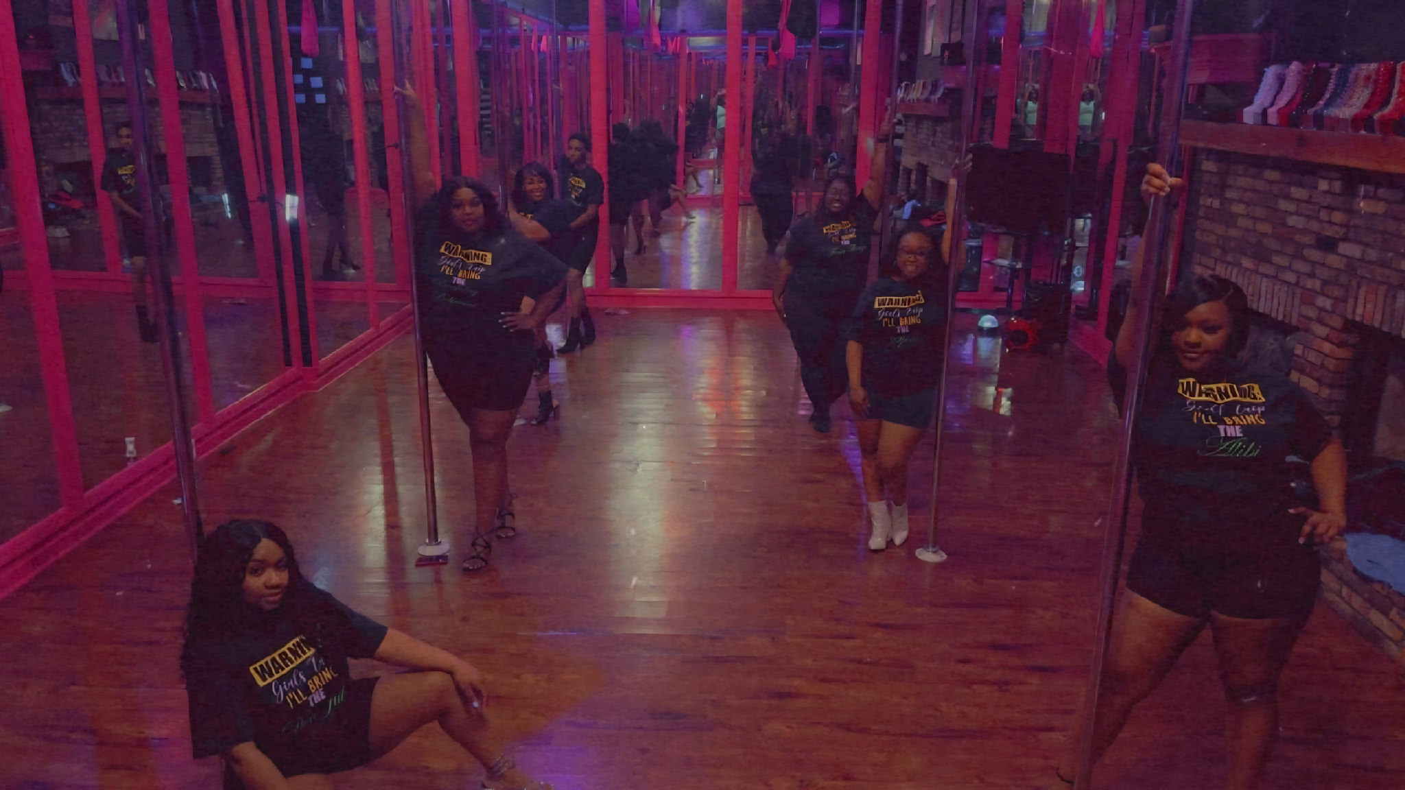 Group of women in black shirts with yellow and green text posing in front of mirrors at a dance studio with brick walls and wooden floor.