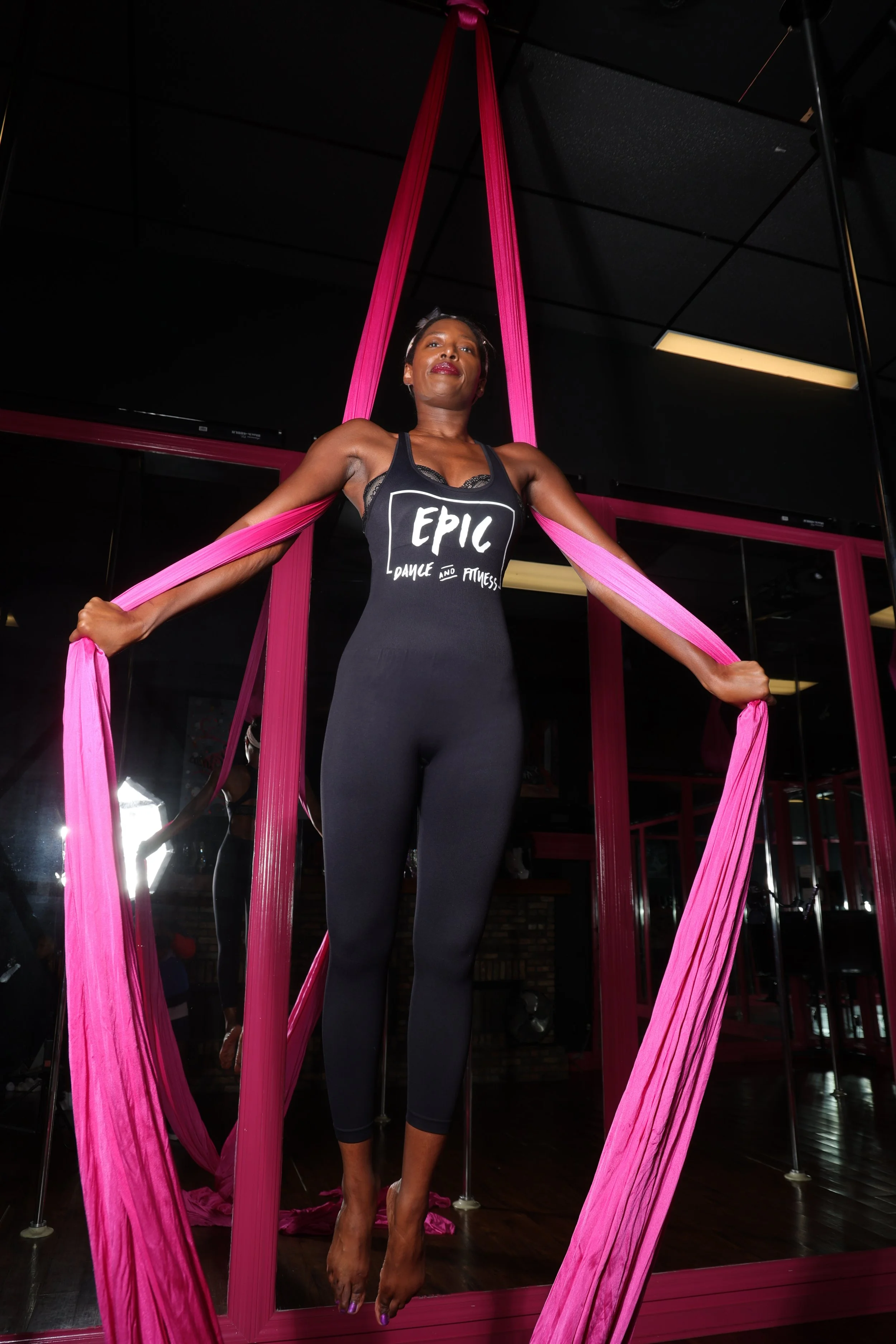 A woman practicing aerial silk fitness in a gym, standing on the floor with pink silks hanging from the ceiling, wearing a black tank top that reads 'EPIC Dance and Fitness,' and black leggings, with a mirror and gym equipment visible in the background.