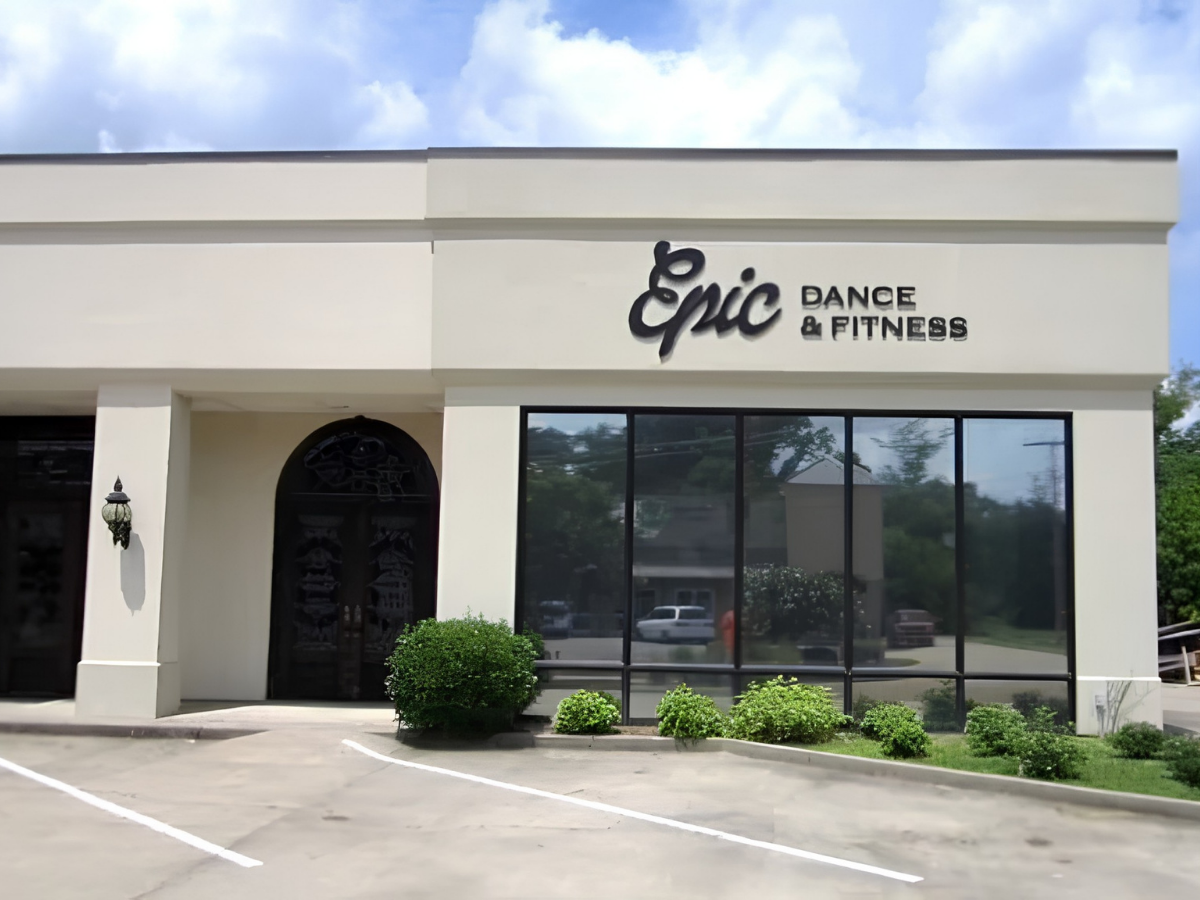 Front view of a white building with a sign that says 'Epic Dance & Fitness,' large windows, and landscaping with bushes and parked cars reflected in the windows.