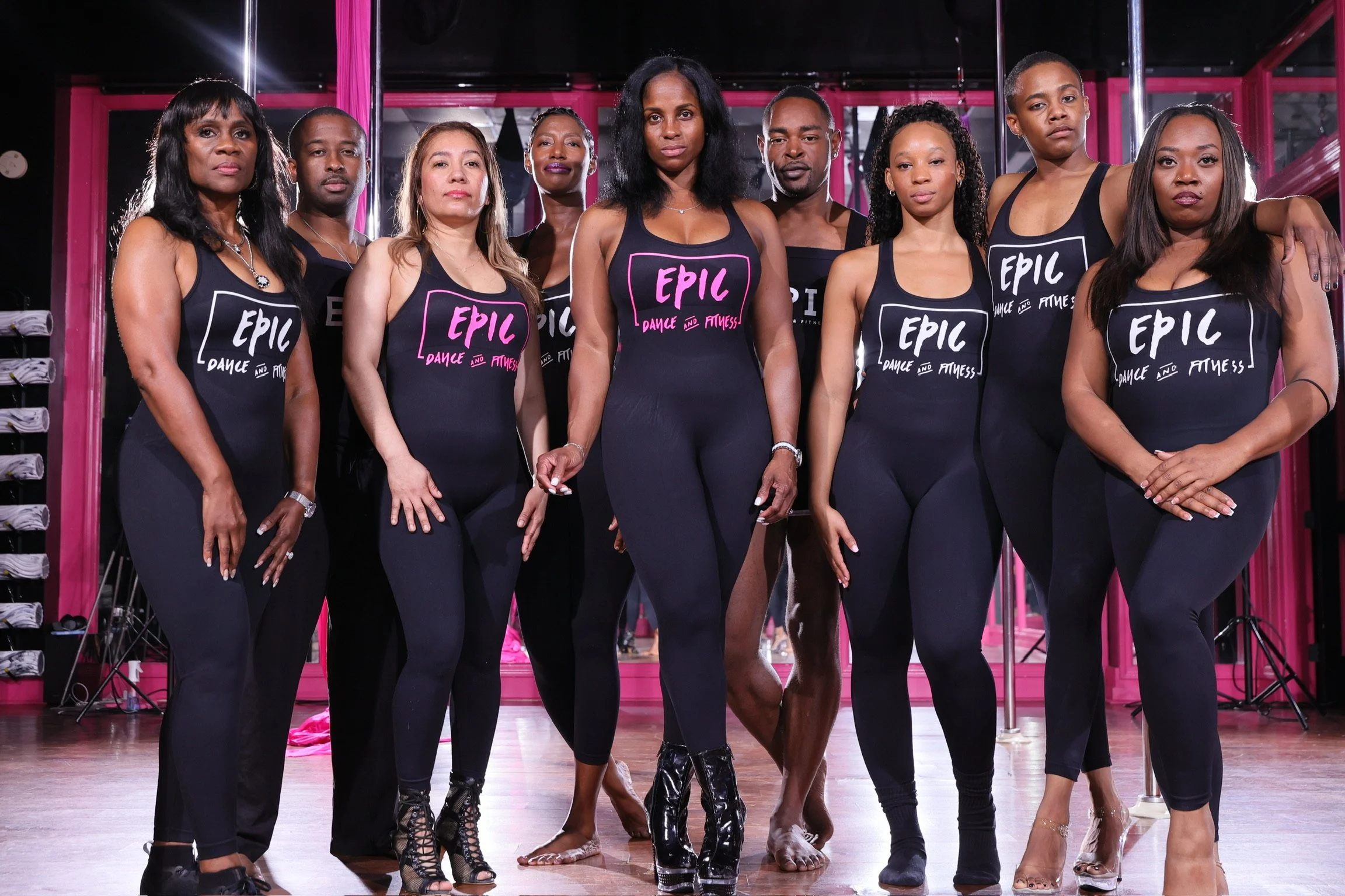 Group of diverse women standing together in a dance studio, wearing matching black tank tops with pink and white text that says 'EPIC Dance and Fitness', posing confidently for a photo.