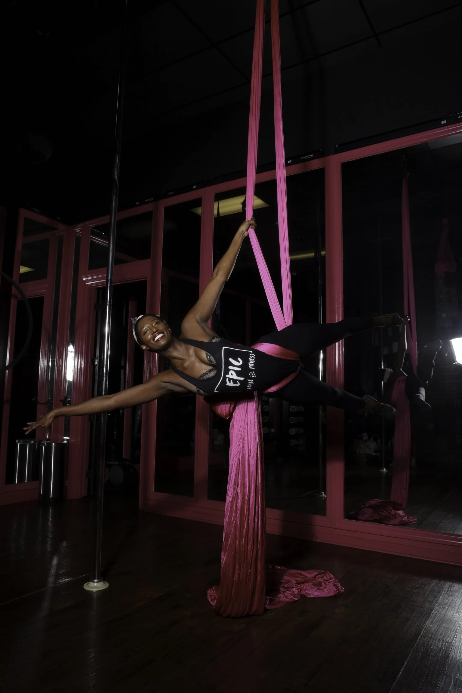 A woman practicing aerial silk yoga in a fitness studio with black walls, a wooden floor, and pink silk fabric hanging from the ceiling.