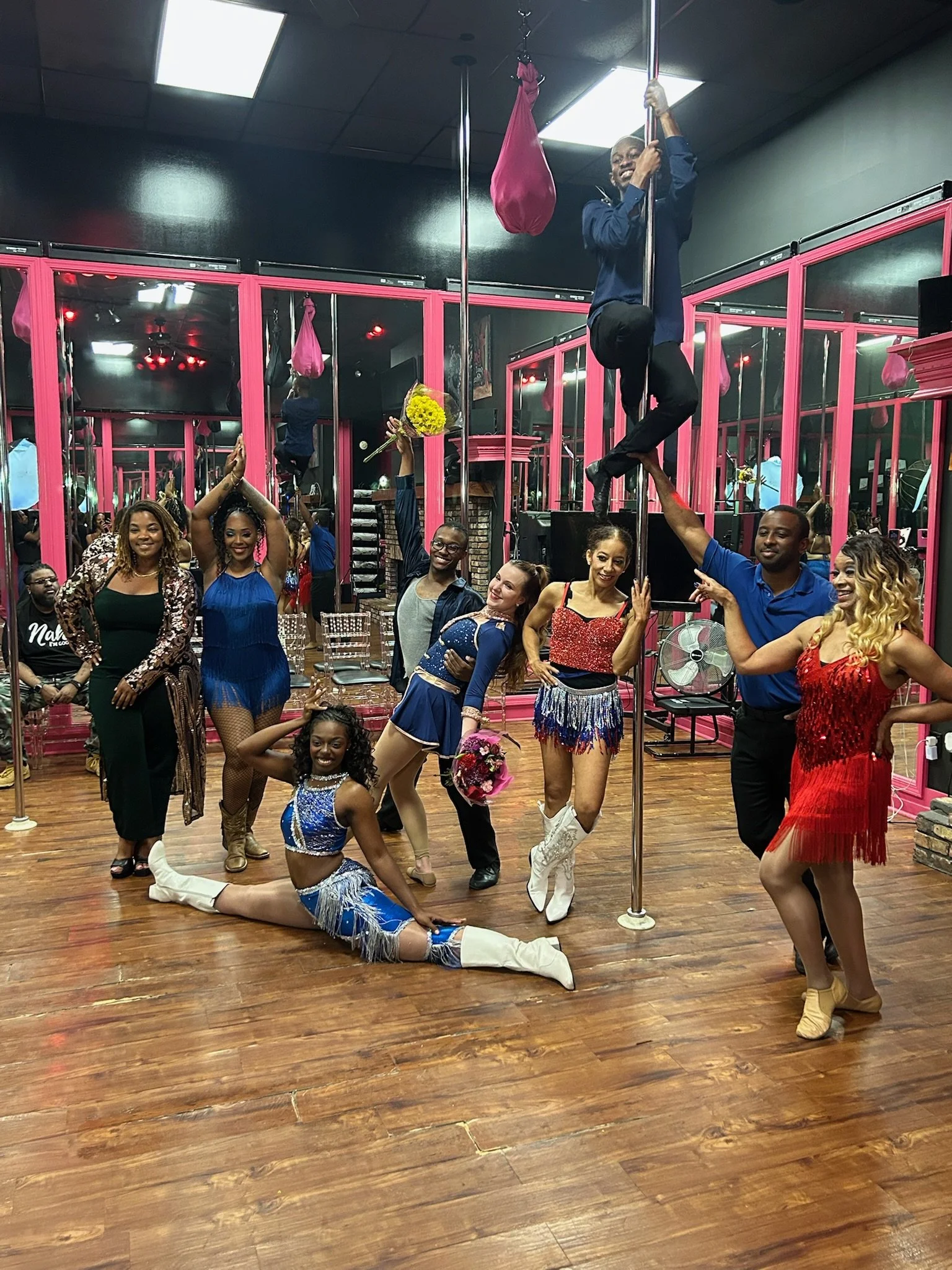 A group of people at a dance or celebration event in a room with pink mirrored walls and wooden floors. Some individuals are dressed in vibrant, fringed costumes, and there is a man on a pole with a pink bag hanging from the ceiling, smiling and holding onto the pole. The group appears to be enjoying a performance or dance.
