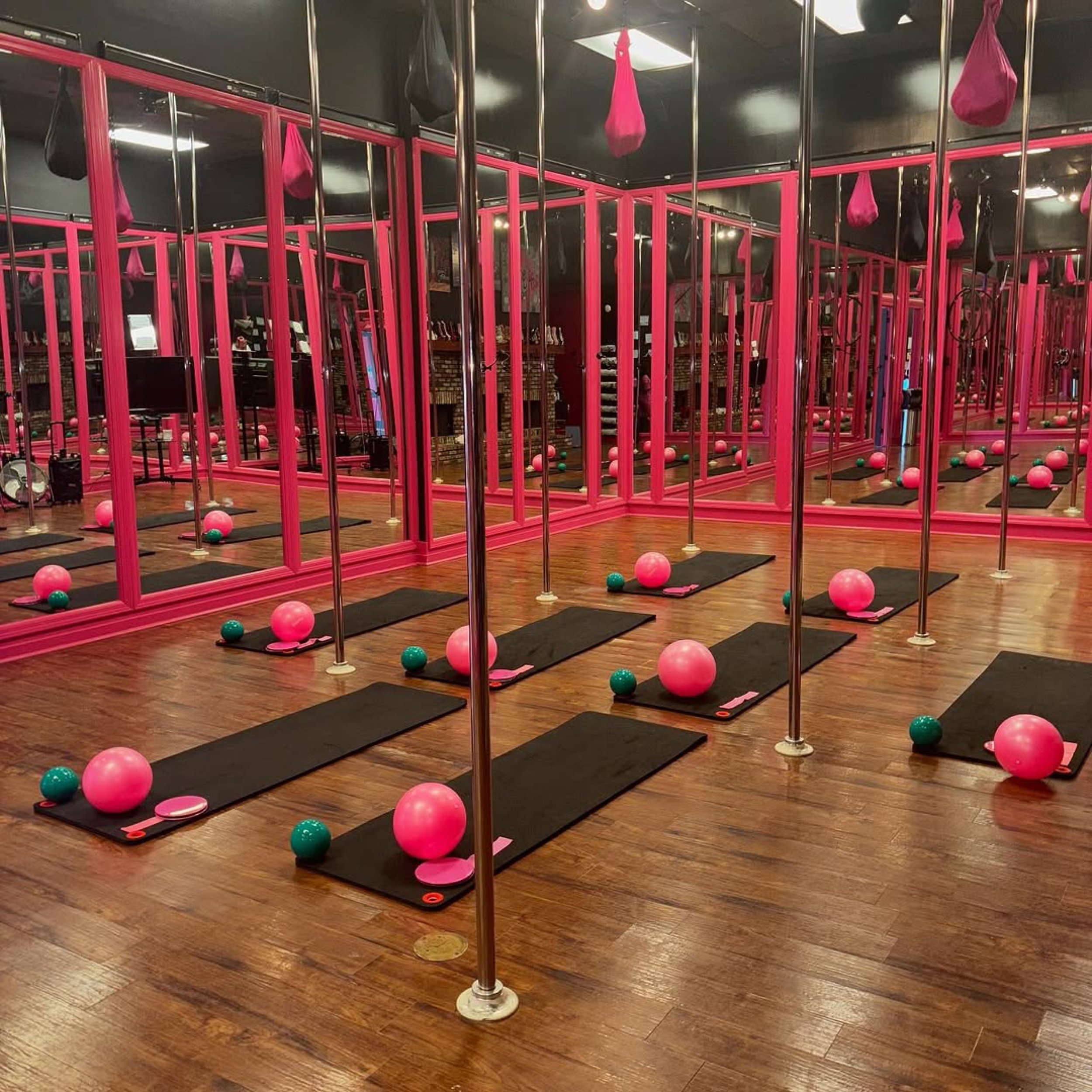 Gym workout area with pink workout cages, pink and green exercise balls, black mats, and metal poles in a fitness studio with wooden floors.