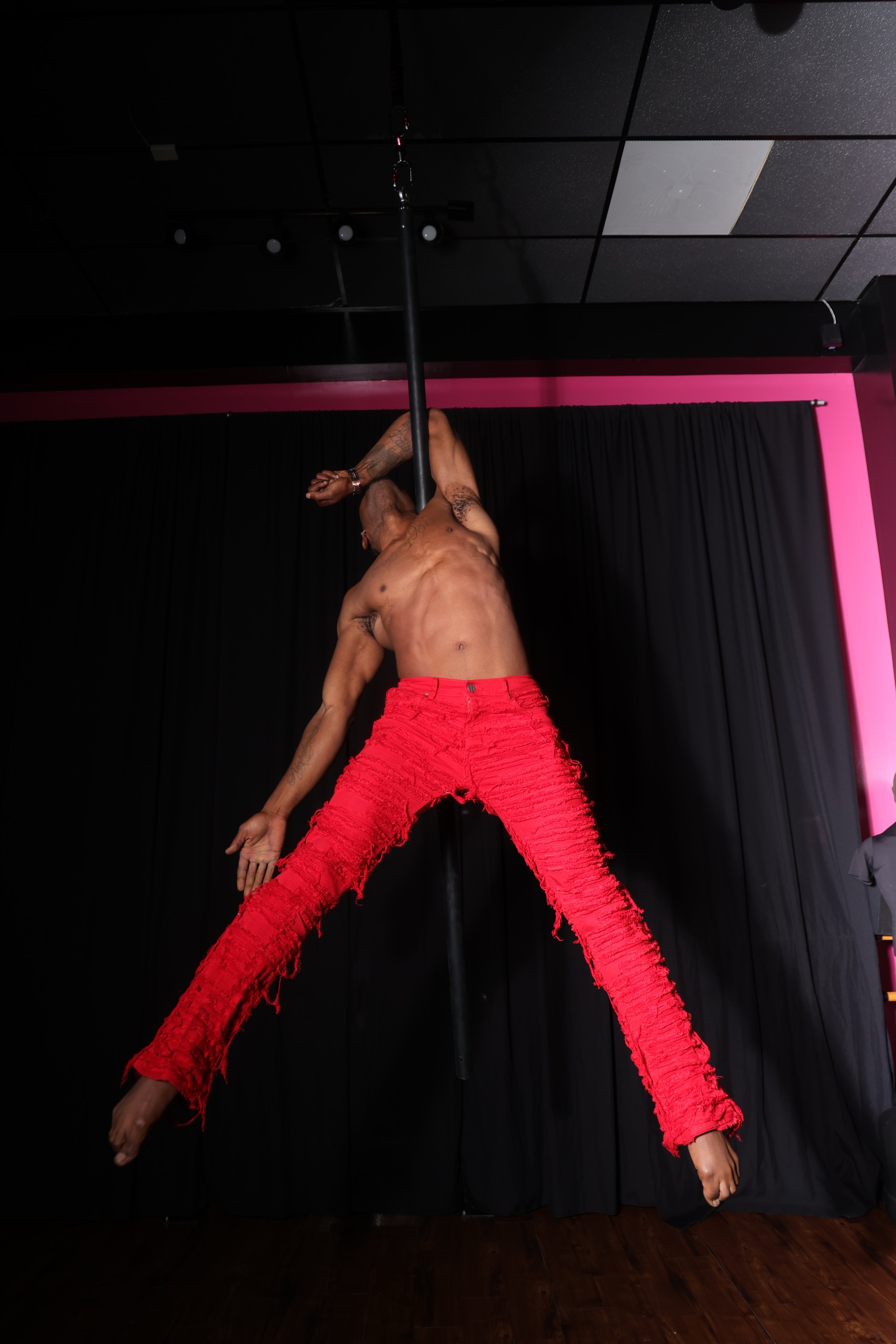 A shirtless man with tattoos on his left arm and chest, wearing bright red distressed pants, is performing a pole dance move on a black stage with black curtains and a pink wall in the background.
