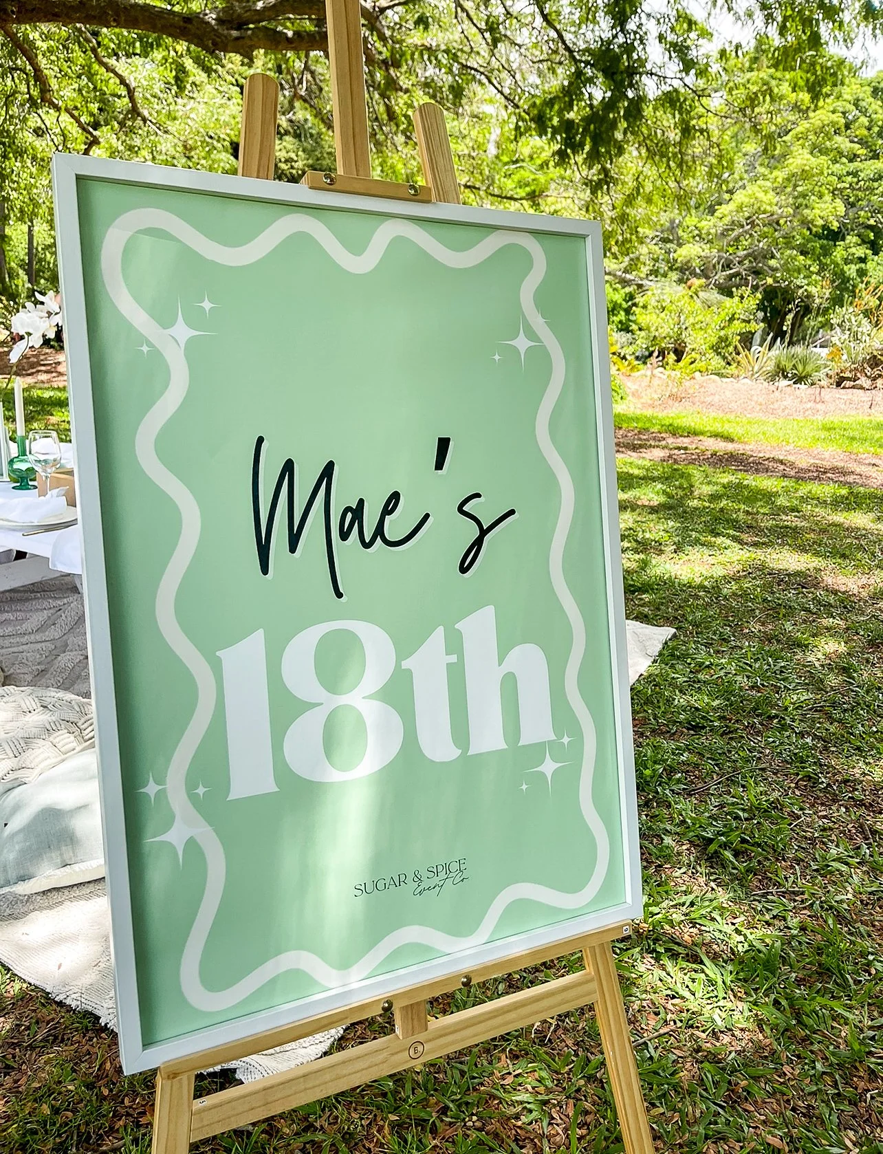 A green celebration sign on an easel reads "Mae's 18th" with a small text "SUGAR & SPICE event Co" at the bottom, set outdoors with trees and grass in the background.