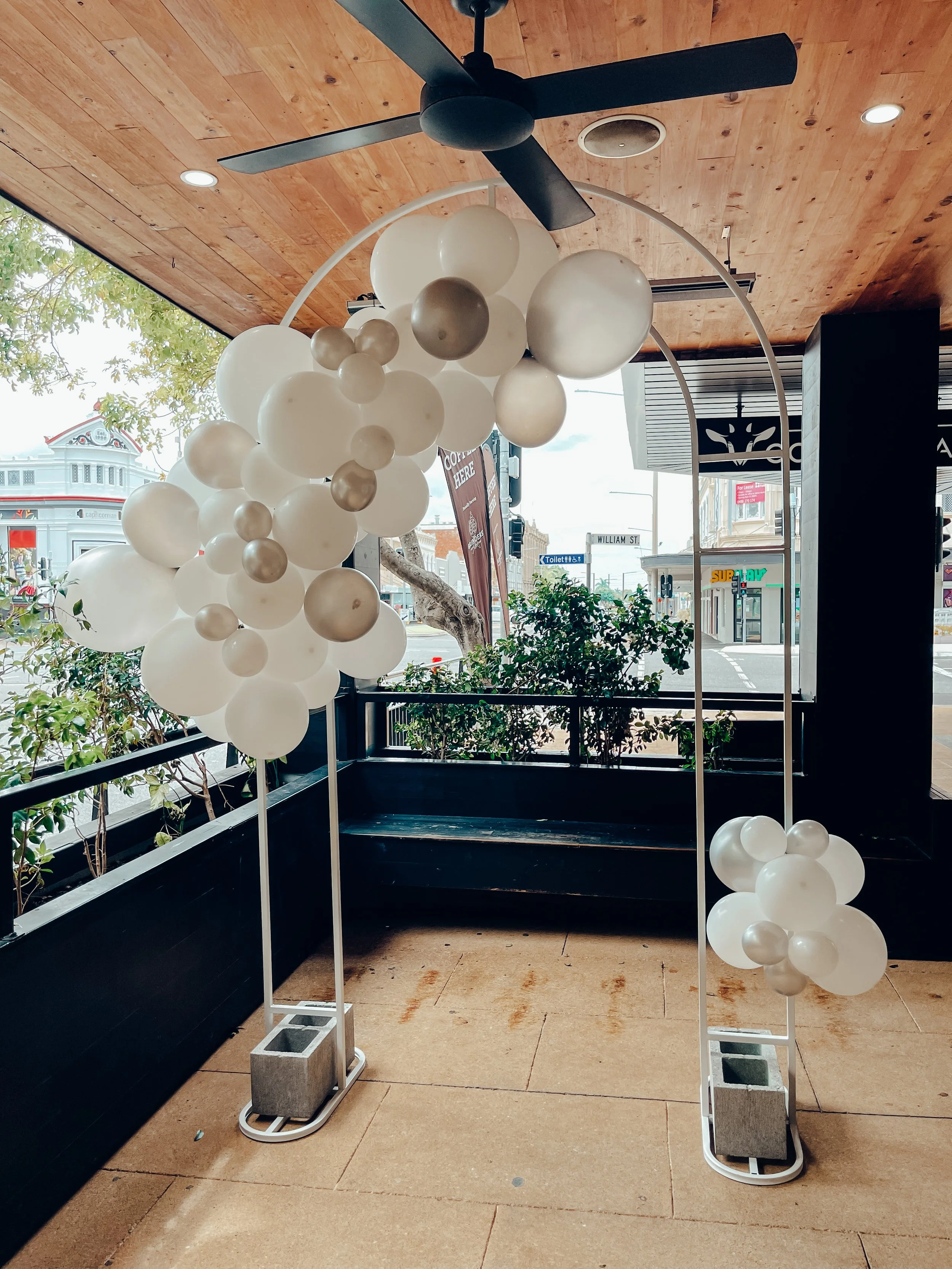 Decorative balloon arch with white and metallic balloons inside a restaurant or cafe, near a window showing street view.