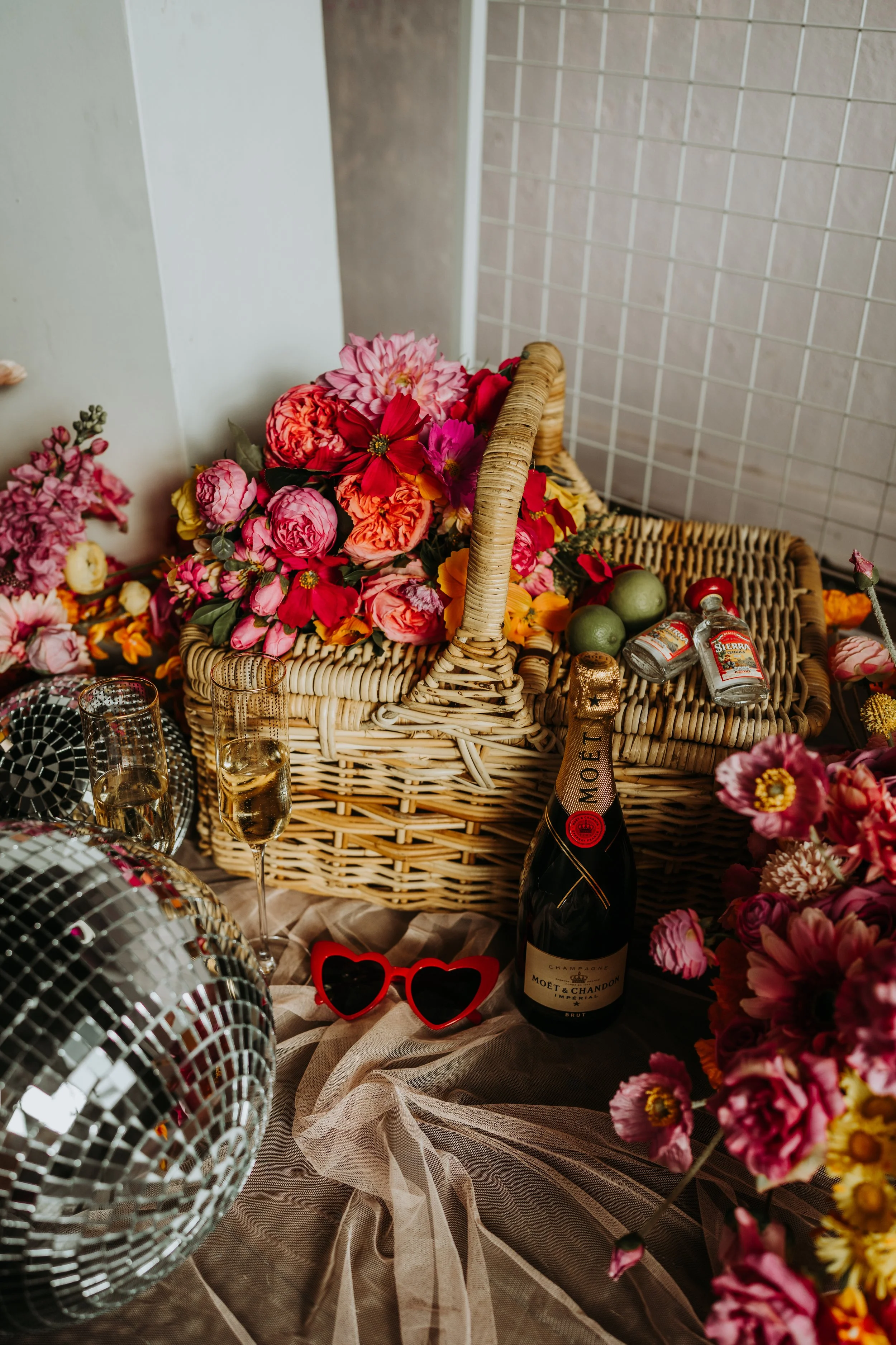 A table decorated with colorful flowers, a bottle of Moët & Chandon champagne, glasses of champagne, a disco ball, and red heart-shaped sunglasses.