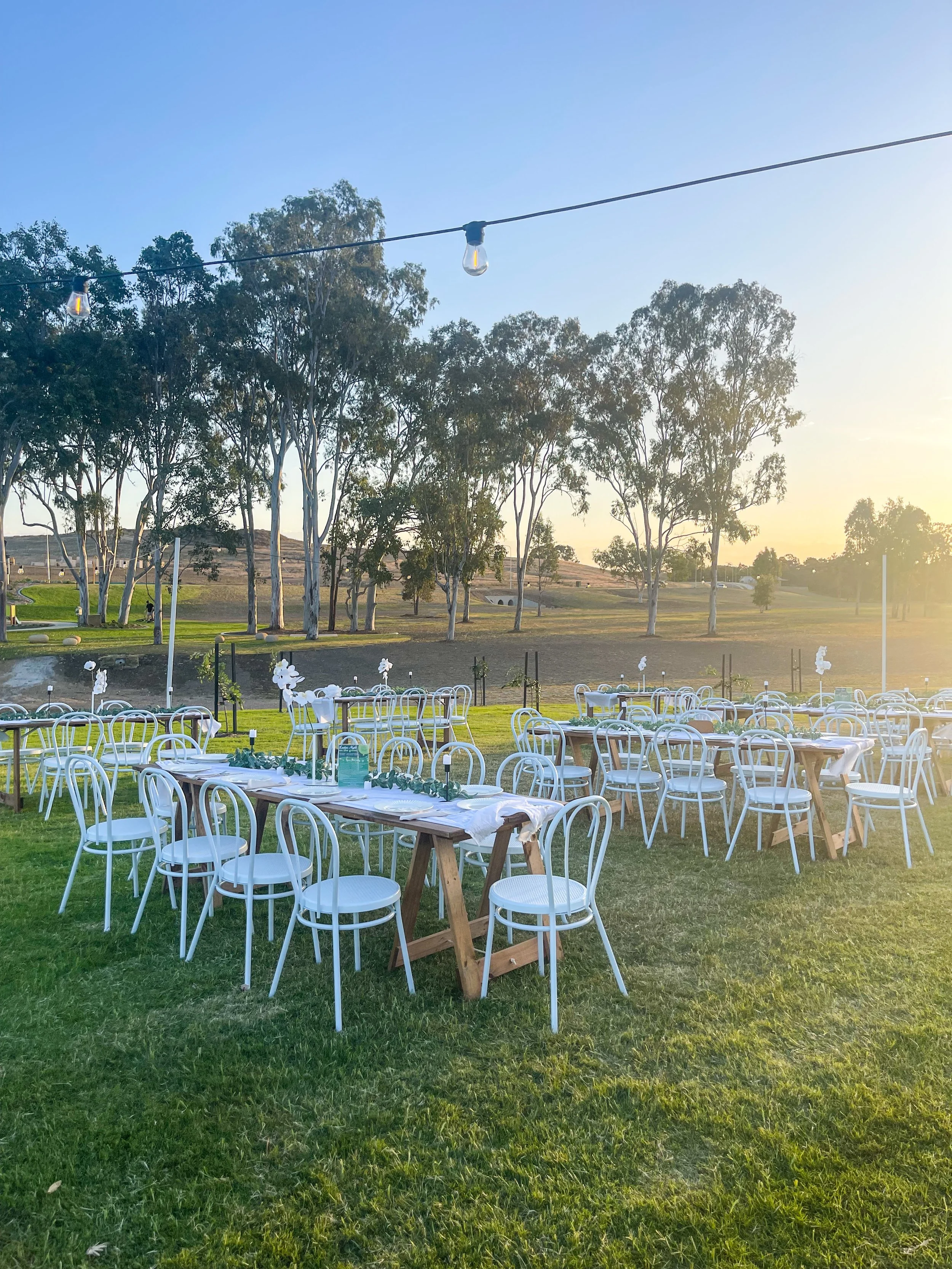 Outdoor event setup with white chairs and tables under a string of lights at sunset, on a grassy field with trees in the background.