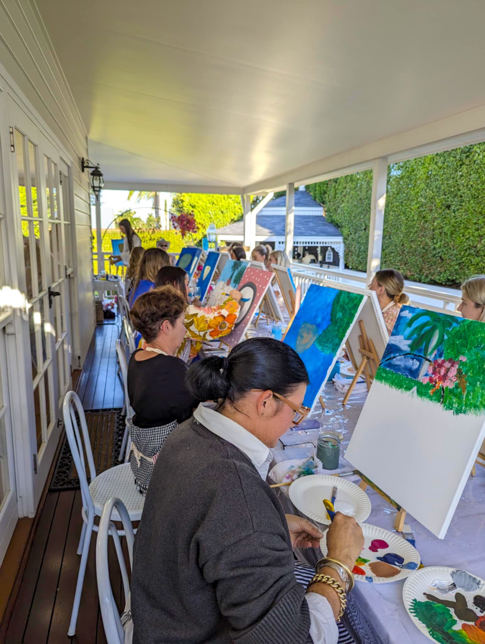 People participating in a group painting class on a covered outdoor porch with easels and canvases, surrounded by greenery.