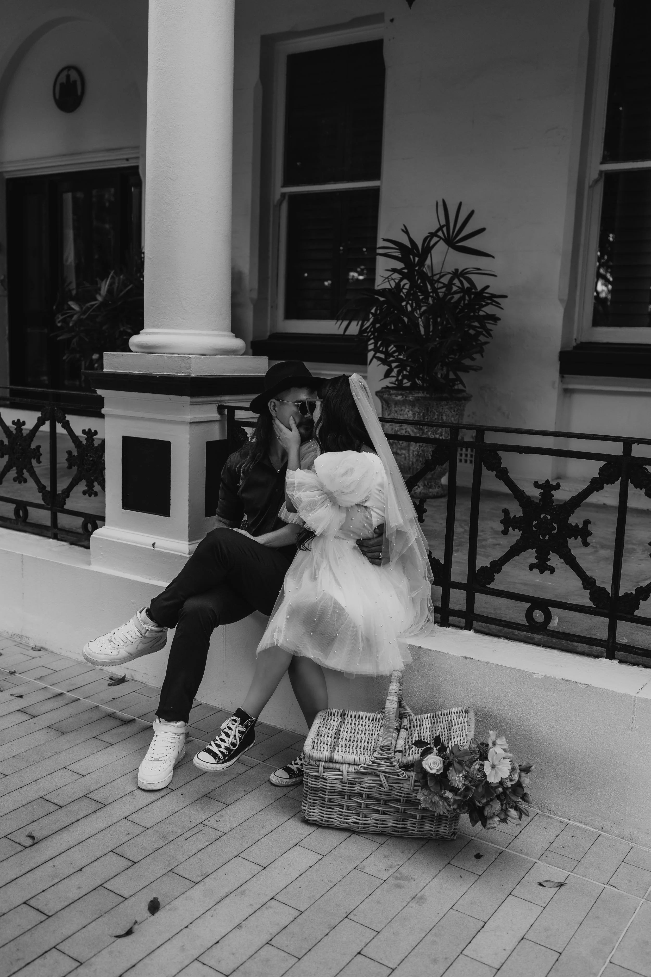 A couple sitting on a ledge, sharing an intimate moment. The woman is dressed in a light dress with a veil, and the man is wearing dark clothing, sneakers, and a hat. There is a basket with flowers on the ground beside them, and a potted plant behind them.
