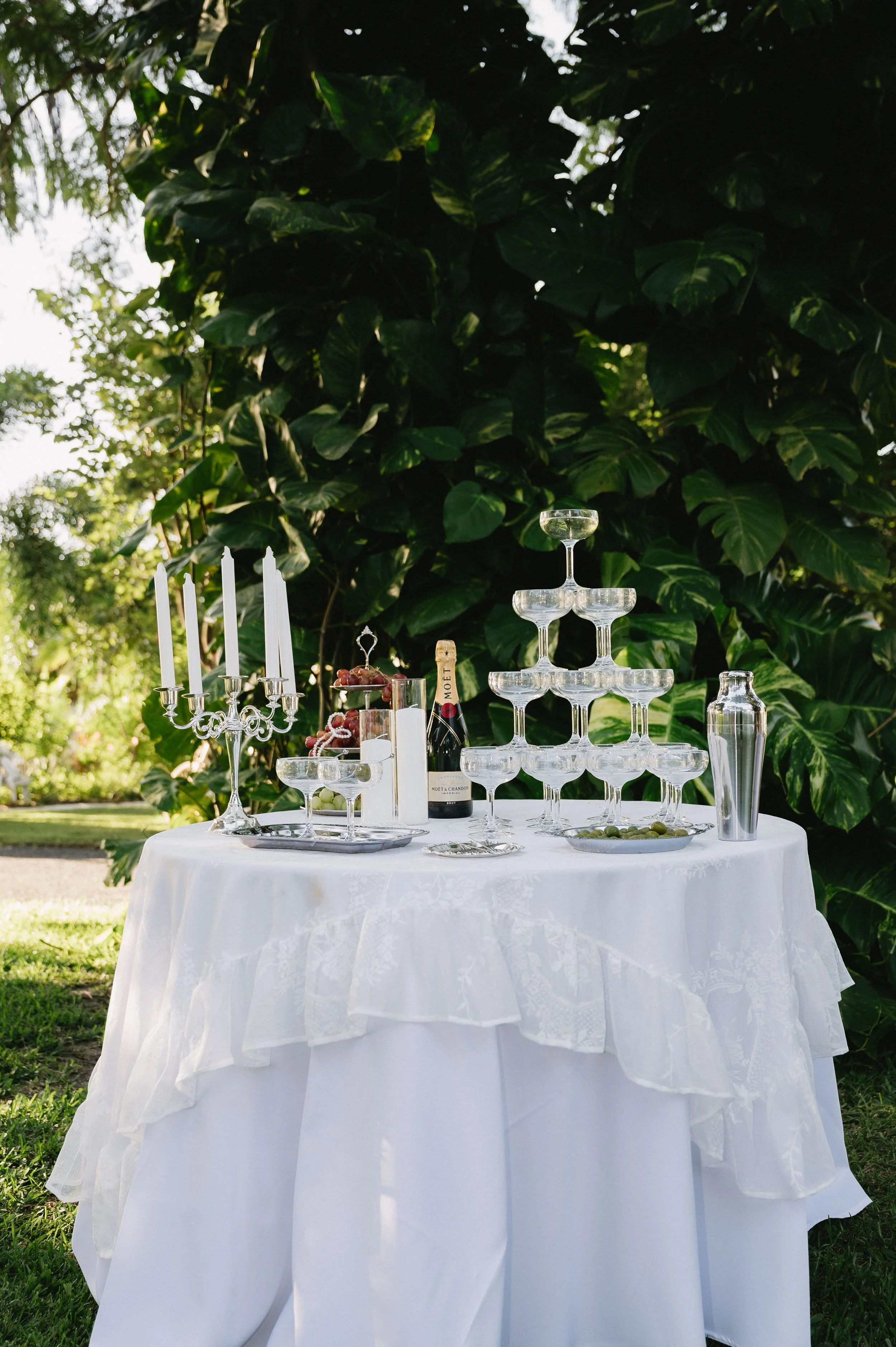 Empty outdoor table covered with a white tablecloth, set for a celebration with a champagne bottle, champagne glasses arranged in a pyramid, a candelabra with white candles, and decorative plates, with lush green foliage in the background.