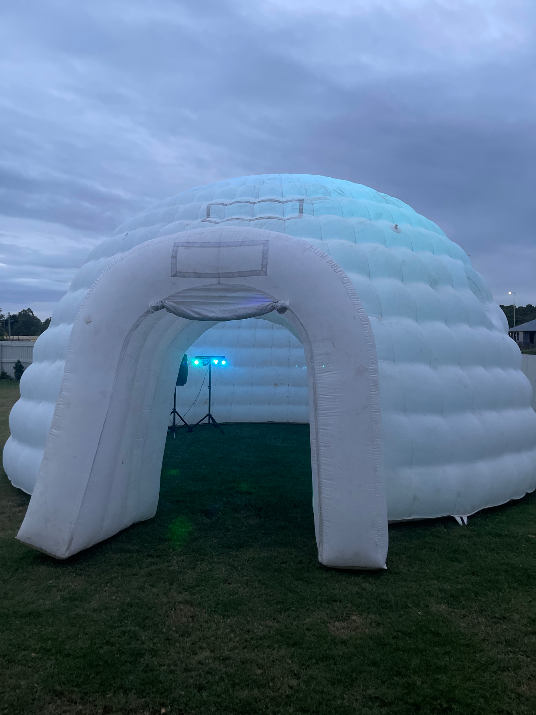 A white inflatable structure resembling a snow cave or igloo is set up on a grassy area during dusk, with a small electronic device emitting blue and green light nearby.